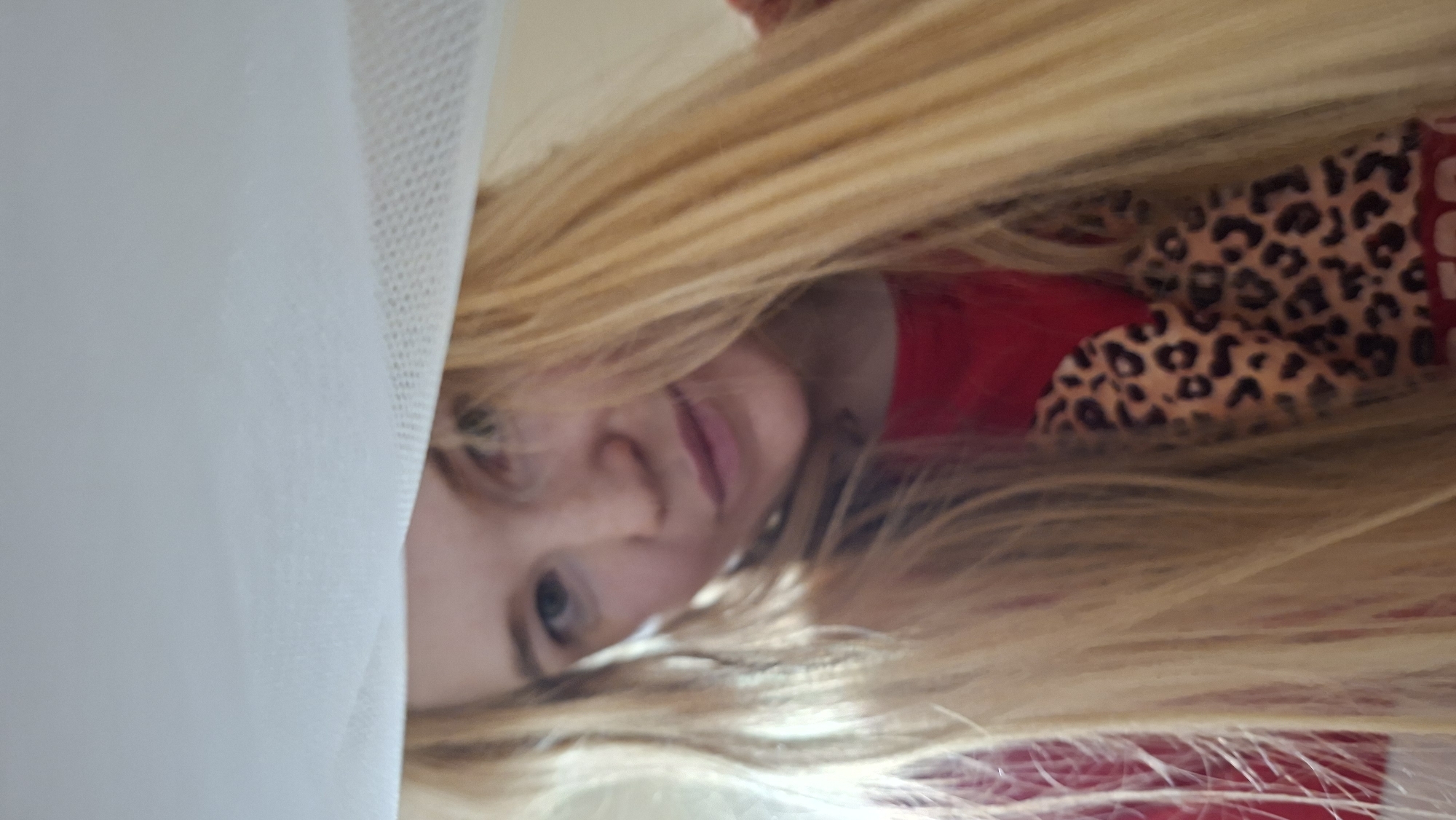 A woman with long blonde hair is looking at the camera, partially obscured by a white cloth. She is wearing a red shirt with a leopard print design on the sleeve. The image appears to be a selfie taken indoors. There is no discernible information within the image to suggest a specific location in Bolshoy Bukor, Russia.