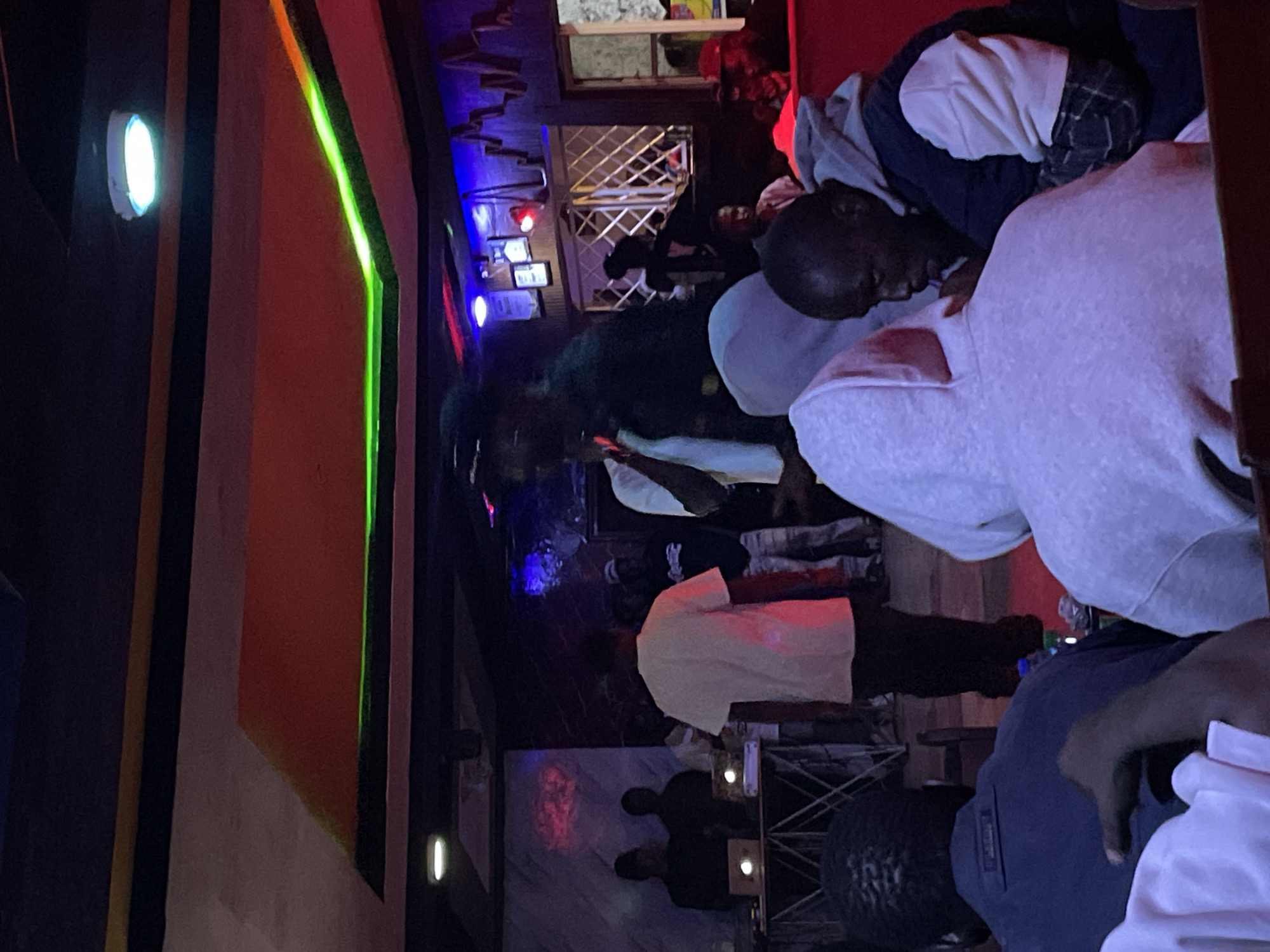 The image depicts an indoor gathering at a dimly lit venue, possibly a club or a performance space, located in Durumi, Nigeria. The environment features colorful lighting, including neon green accents along the ceiling and blue and red lights illuminating the back wall. A stage area is visible in the background with several individuals standing. One person in a white t-shirt appears to be addressing the crowd, possibly as a performer or MC. Another individual in a black hoodie is standing near the stage. In the foreground, a group of people are seated, facing away from the camera. They are wearing casual clothing, including hoodies. The overall atmosphere suggests a lively social event.
