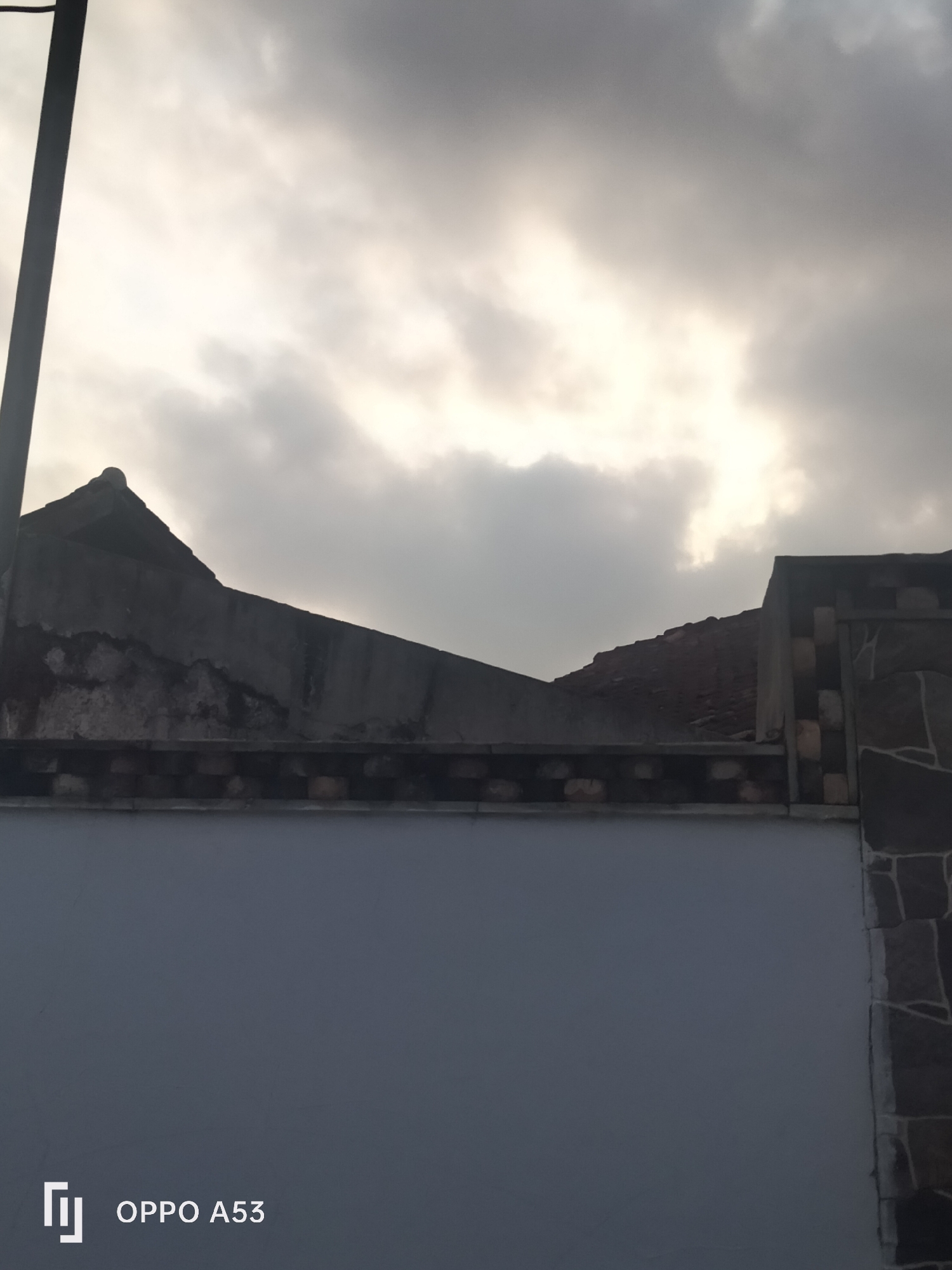 The image captures a cloudy sky above a two-tiered structure in Badung, Indonesia. The top layer consists of a dark, angular rooftop with visible wear and damage, suggesting an older construction. Below this is a decorative band made of alternating dark and light rectangular bricks. The lowest visible element is a smooth, light grey wall, which extends across the bottom of the frame. To the right, a section of the wall is adorned with irregularly shaped, dark brown and grey stones. A dark vertical pole is partially visible on the far left side of the image. The Oppo A53 watermark is present in the lower left corner.