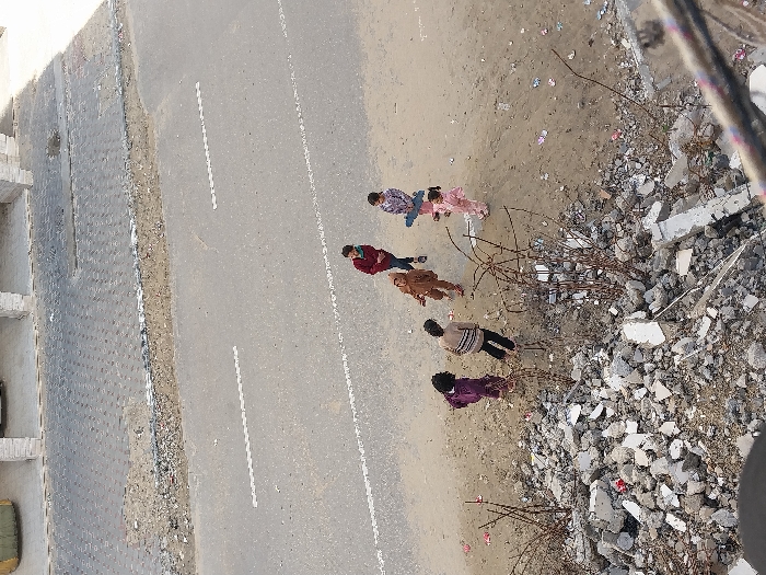 An elevated view captures a street scene in Khan Yunis, Palestinian Territory. In the foreground, a substantial pile of concrete rubble and exposed rebar occupies the left side of the frame, indicating structural damage or demolition. Adjacent to this rubble, a dirt and sand shoulder transitions to an asphalt road with faded white dashed lane markings. Six individuals, appearing to be children, are gathered on this dirt shoulder.
From left to right, the individuals are: one child in a dark purple top and dark pants, another in a horizontally striped top and dark pants, a third in a light brown hooded outfit, a fourth in a dark red top and dark pants, a fifth in a plaid top and light pants holding a light blue rectangular object, and a sixth, smaller child in a pink outfit, positioned slightly apart from the main group. All individuals appear to be looking generally towards the center of their gathering. The dirt shoulder contains scattered small pieces of debris. In the background, beyond the road, a paved area leads to a building featuring multiple light-colored stone pillars. Bright daylight illuminates the scene, casting distinct shadows.