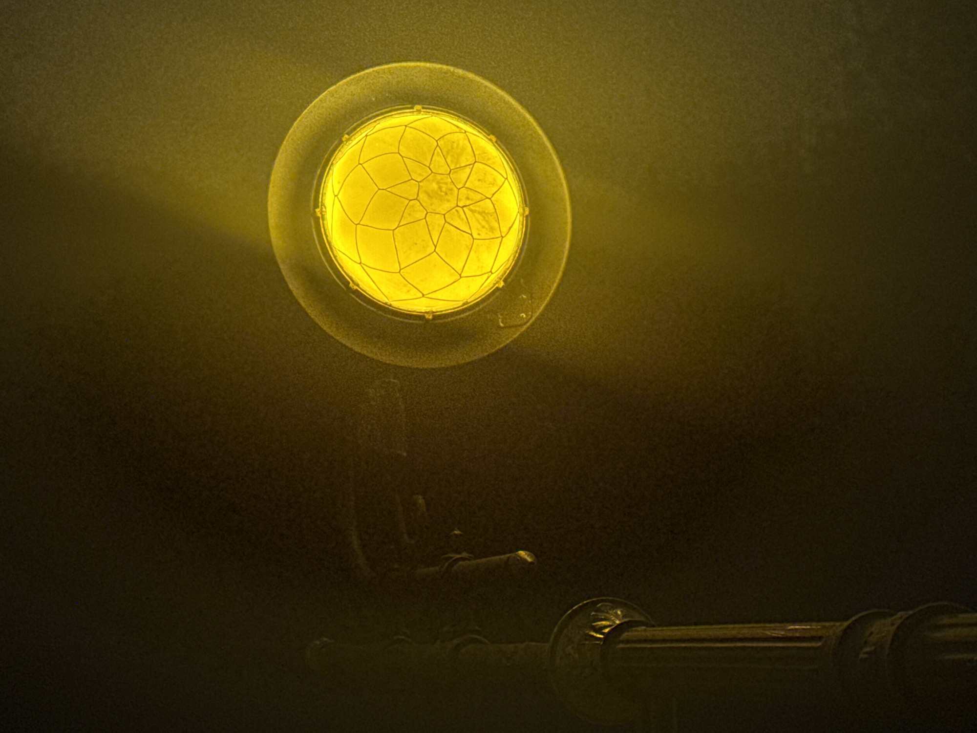 The image presents a prominent, circular light fixture emitting a vibrant yellow glow, serving as the central subject. The light source is encased or covered by a distinctive, intricate web-like pattern, resembling cracked glass or a decorative grill. This strong illumination casts a warm, golden hue across the otherwise dark and textured background, which appears to be a wall or a surface dimly lit by the lamp's output. Below the luminous sphere, obscured in shadow but catching some of the light, are metallic structures. These appear to be pipe-like or decorative architectural elements, possibly made of brass or bronze, showing some ornate detailing, particularly a fluted or sculpted section towards the lower right.
The scene conveys a sense of antiquity and mystery, characteristic of an old interior, possibly within a historical building or industrial setting. The intense yellow light creates an atmospheric effect, suggesting either an old-fashioned lamp or a specific type of warning light. The background's grainy texture might indicate dust in the air or a rough surface, further enhancing the aged ambiance. There are no people visible, and no specific event is occurring; the image is a still life focusing on the interplay of light and shadow on architectural elements. The setting is clearly dark, relying entirely on the artificial light source. This scene could be found in an older, atmospheric location within Saint Petersburg, Russia. No visible text is present in the image.