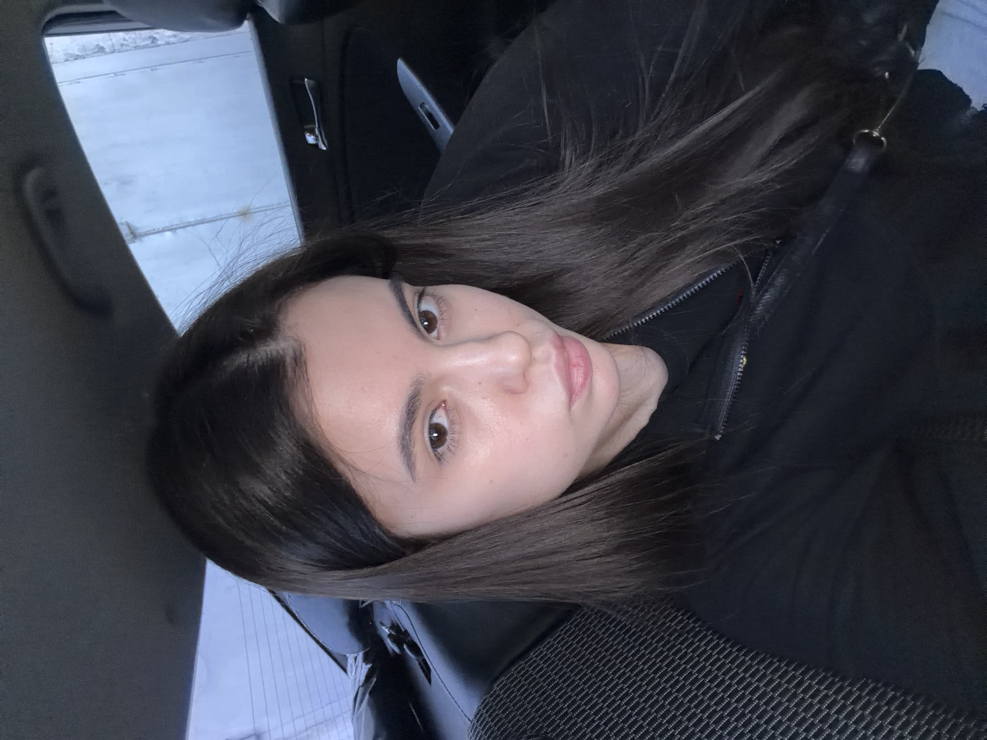 A young woman with long, dark brown hair is in the driver's seat of a vehicle. She is wearing a black jacket. The interior of the vehicle is visible, including a black seat with a textured pattern. Outside the window, there is a view of a structure with a metallic exterior. This is in the city of Chaikovsky, Russia.