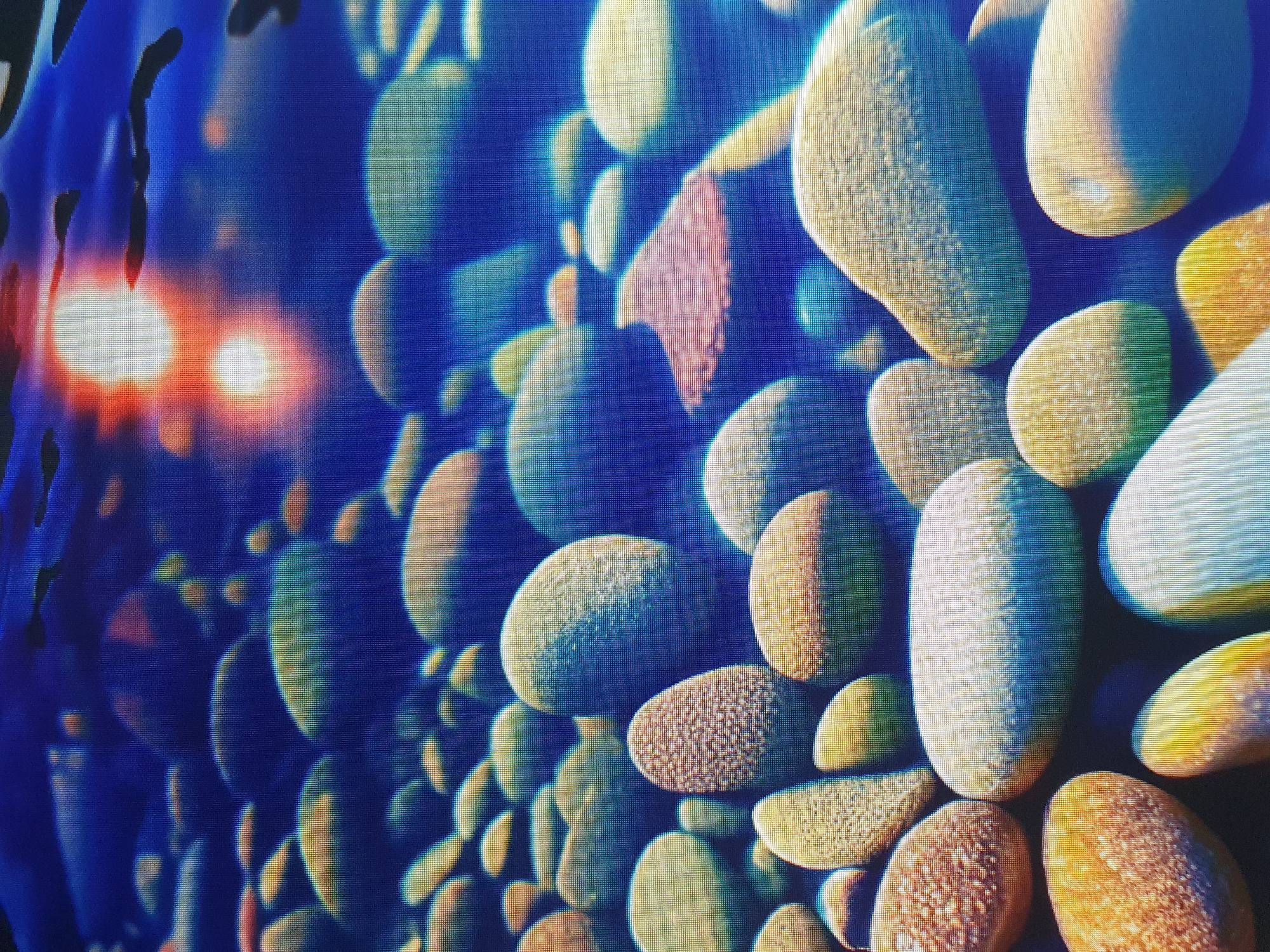 The image depicts a close-up, underwater view of smooth, rounded pebbles and stones of various sizes and colors, primarily in shades of blue, yellow, and white, with some reddish-brown accents. The stones are densely packed and appear to be submerged in water, as indicated by the rippling surface and a bright, blurred light source resembling the sun or a light reflection near the top of the frame. The scene is entirely devoid of any human presence or activity. The image does not provide any information that can be used to identify a specific location in the city of Tashkent, Uzbekistan, or any other city for that matter. It is a generic depiction of submerged stones.