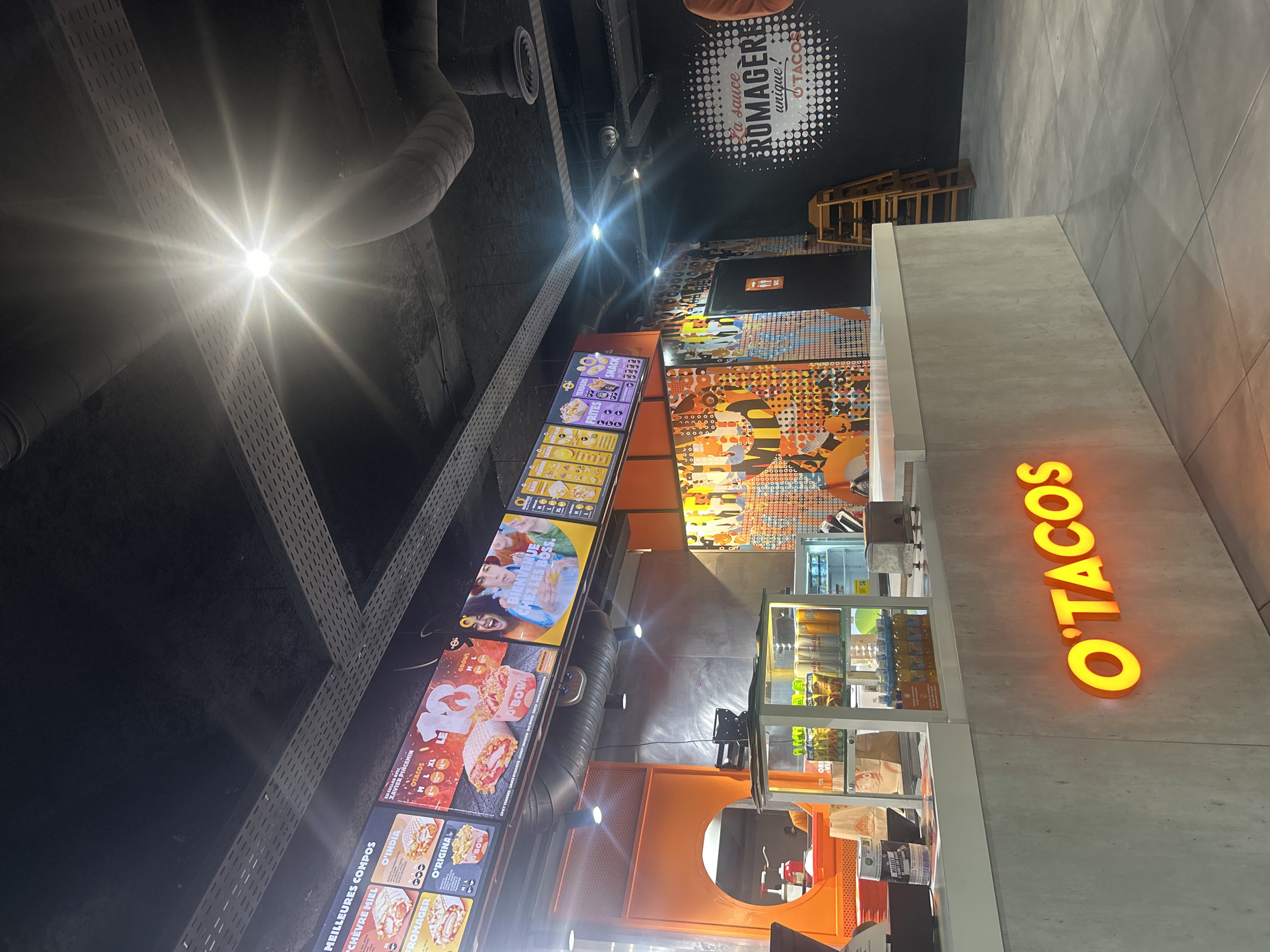 This indoor shot showcases an "O'TACOS" restaurant, likely located in Champs-sur-Marne, France, based on the French text on the menu. The restaurant features a vibrant orange and grey color scheme. A bright light fixture illuminates the scene from the upper left corner. A menu board displays various food offerings with French descriptions. The ordering counter features the "O'TACOS" logo in illuminated letters. A refrigerator displays drinks, and a service window allows customers to see inside the kitchen area. A wall graphic with the phrase "La sauce ROMAGERE unique! d'TACOS" is visible in the upper part of the image. In the background, there are some ladders.