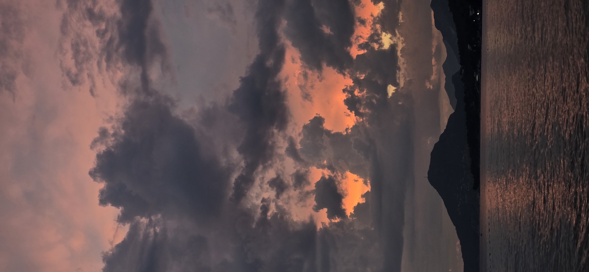 The image captures a dramatic sunset over the sea in Bar, Montenegro. Silhouetted against the vibrant, cloud-filled sky are the dark forms of mountains and hills. The water in the foreground reflects the fiery hues of the sunset, creating a shimmering, textured surface. Scattered buoys are visible on the water, marking a protected area or swimming zone. The sky is a spectacle of color, with deep grays and blacks of heavy clouds interspersed with brilliant oranges and pinks where the sun's rays are breaking through. The overall impression is one of natural beauty and a serene end to the day.