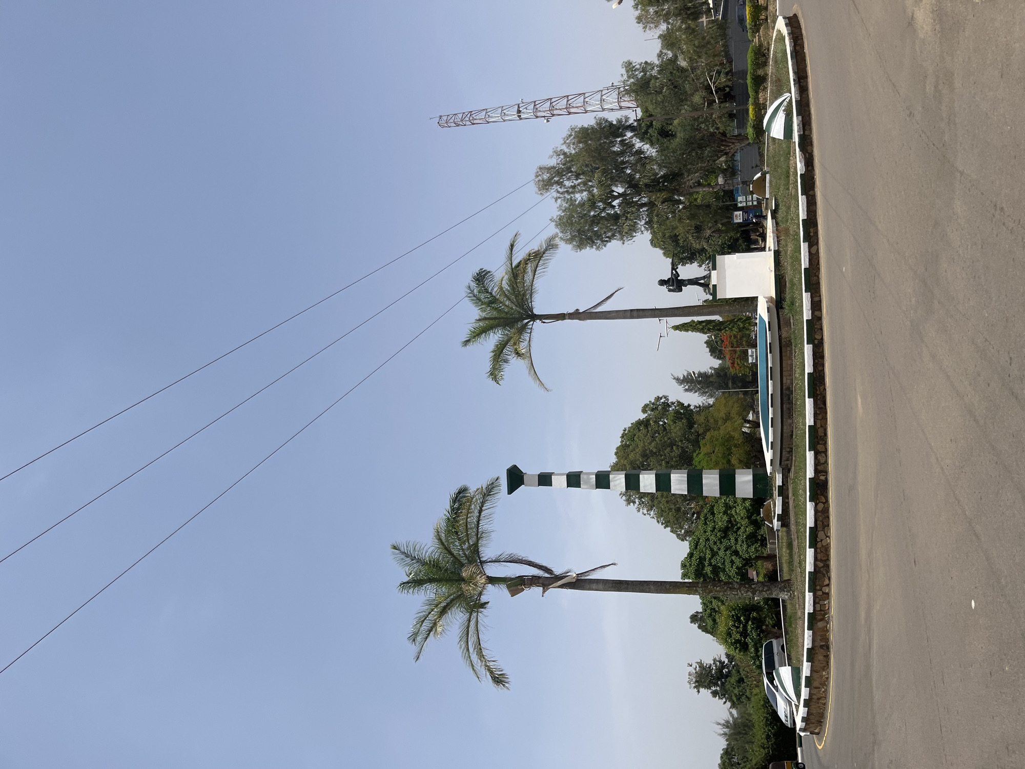 The image depicts a street scene in Jos, Nigeria, notable for a roundabout featuring two palm trees and a striped pole, likely a landmark or decorative feature. A monument with a statue is also visible in the background. Overhead electrical wires traverse the sky, and a communications tower stands in the distance. The road surface is paved, and the vegetation appears lush, indicative of a tropical or subtropical climate.