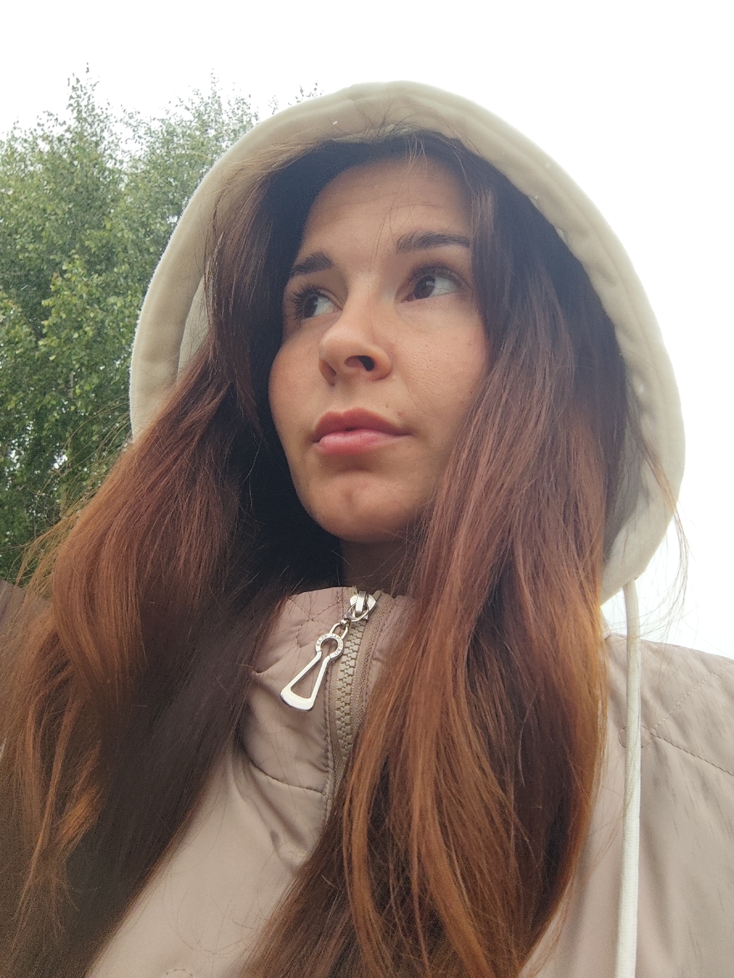 A close-up, low-angle shot shows a young woman with long, auburn hair wearing a beige hooded jacket. She is looking upwards and to her left, with her mouth slightly open. The background consists of a blurry green tree and a white sky. The image appears to be taken outdoors in the city of Fokino, Russia.
