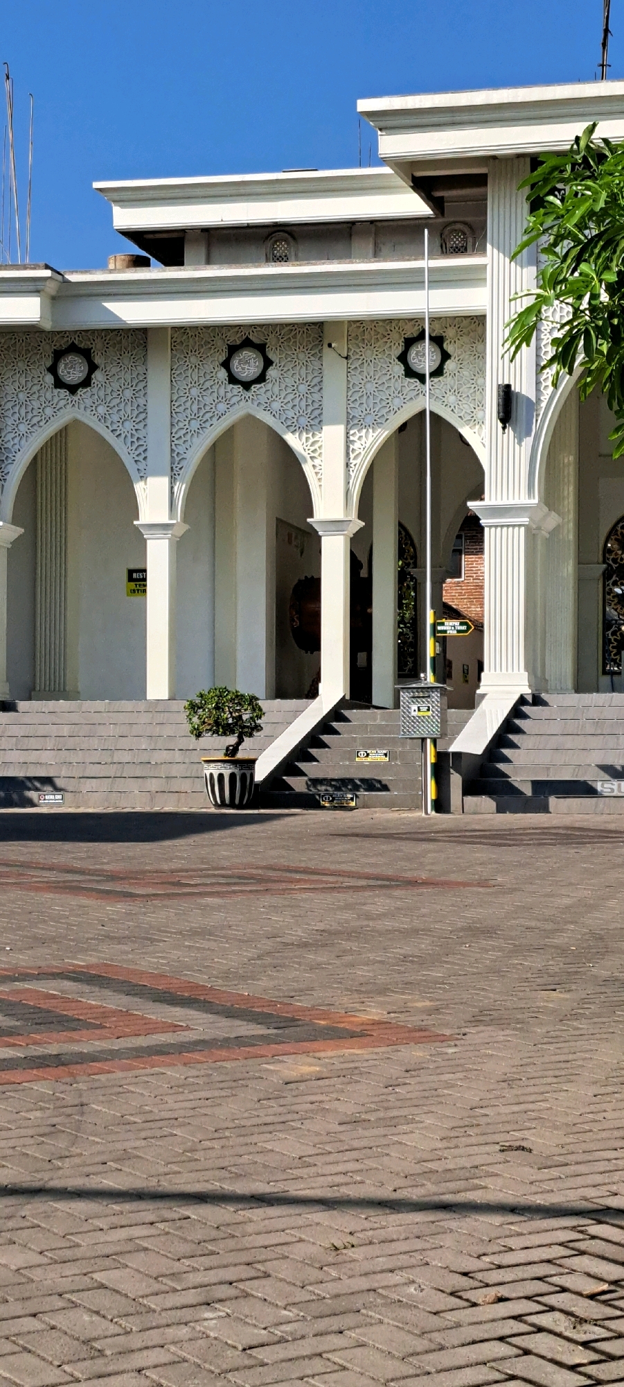 The image depicts the entrance to a building, likely a mosque, in Sumber Gedang, Indonesia. The architecture is characterized by white columns, arched doorways with intricate Islamic geometric patterns, and a tiled stairway leading up to the entrance. A single potted bonsai tree sits on the left side of the stairs, adding a touch of greenery. The ground in front of the building is paved with interlocking bricks, with a pattern of red and gray stripes. The sky is clear and blue, suggesting a sunny day. There are no people visible in the frame. The overall impression is one of a well-maintained and serene public space.