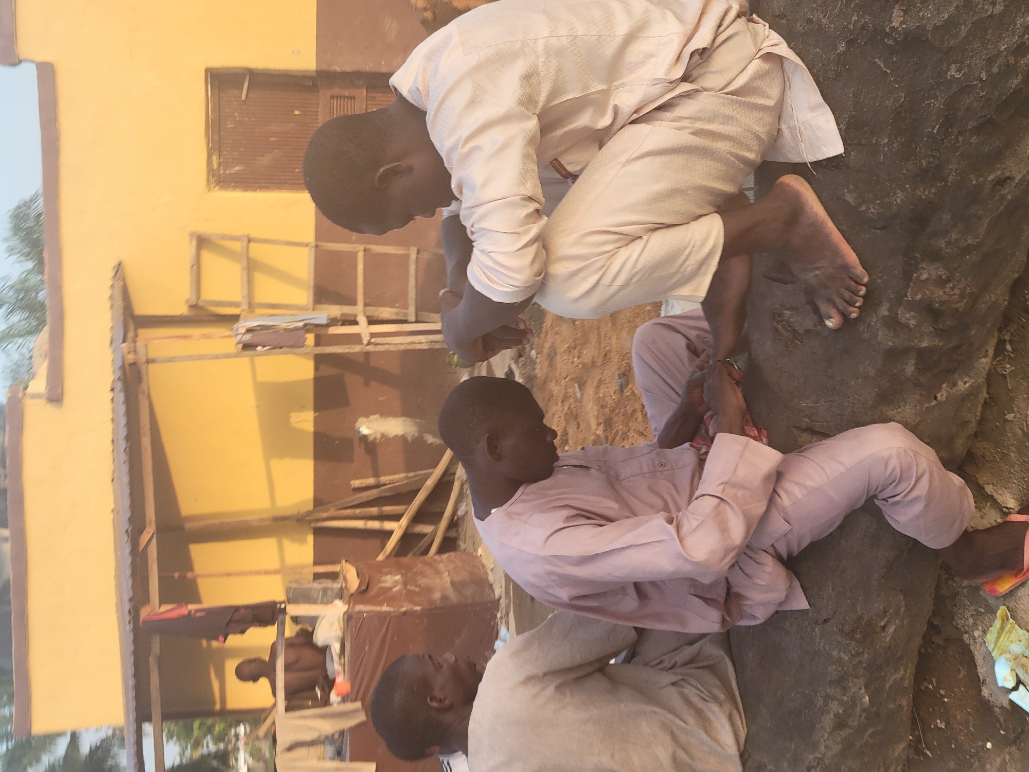 Three African males are present in an outdoor setting near a building in Kwamba, Nigeria. One man is crouched on a large, dark rock with bare feet, wearing a light-colored, long-sleeved outfit. A second man sits on the rock, clad in a pinkish-purple, long-sleeved ensemble. A third man is partially visible, kneeling and wearing a light-colored outfit. A building with a mustard-yellow exterior is in the background, featuring a boarded-up window and wooden scaffolding.