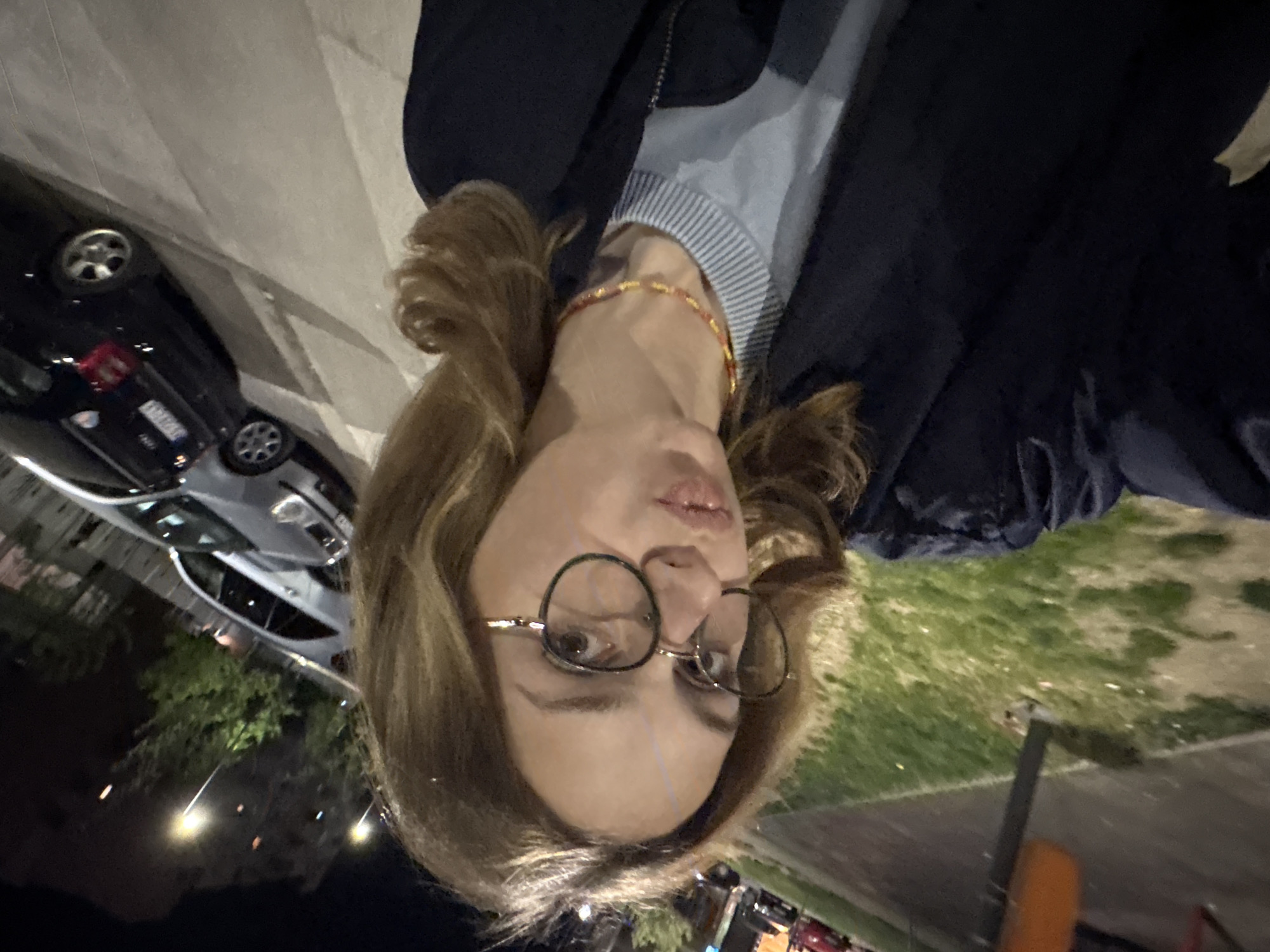 An outdoor, nighttime image features a person, likely a woman, with light brown hair and round, dark-framed glasses, in the foreground. She is looking towards the camera with a slightly parted mouth and a neutral to pensive expression. She wears a light blue and grey striped top under a dark blue or black jacket, and a small, beaded bracelet with red and gold elements on her wrist is visible.
The background reveals an urban or suburban setting at night. Several cars are parked or driving; a dark-colored sedan with a partially visible Serbian license plate "KG 030-C" is prominent on the left, confirming the location as Kragujevac, Serbia. Another lighter-colored car is behind it. Streetlights illuminate the scene with warm, yellow light, casting a glow on patches of green grass or foliage. Buildings are faintly visible in the distance. The overall atmosphere is calm and quiet, typical of a city at night.