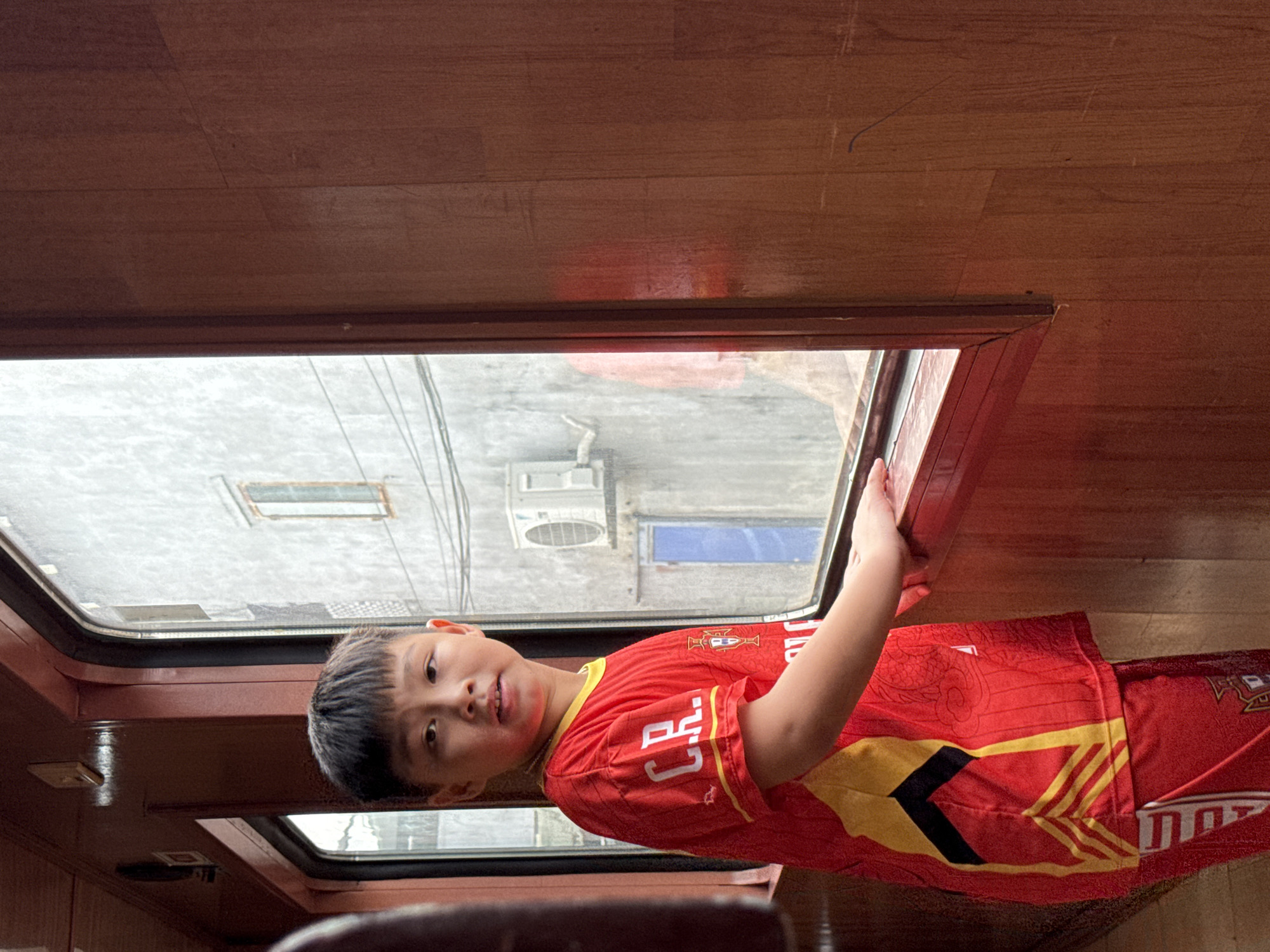 An East Asian boy with short black hair stands inside a train car, looking out a window. He is wearing a red and yellow soccer jersey with the letters "C.R." on the left sleeve and "CR7" across the front. The boy's right hand rests on the window sill. The train car interior is paneled with dark wood. Outside the window, a weathered, grey building with an air conditioning unit and a blue door is visible. The scene is set in Da Nang, Vietnam.