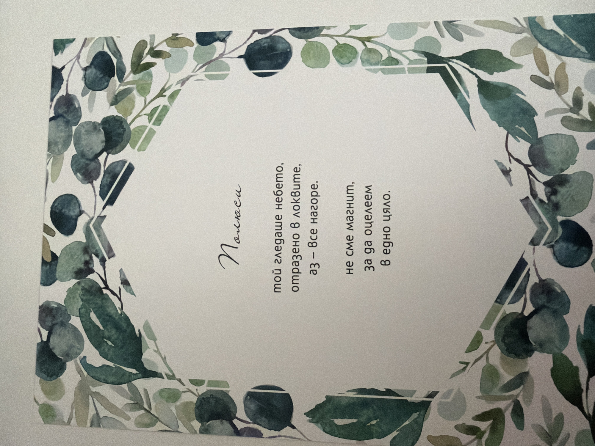 The image is a close-up shot of a card with a watercolor floral border. The card contains text in Bulgarian. The top of the card has a title in a cursive font that reads "Поляна". Below the title, there are two stanzas of poetry. The first stanza reads: "мой гледаше небето, отразено в локвите, аз – все нагоре." The second stanza reads: "не сме магия, за да оцелеем в едно цяло." The card is set against a plain background. There are no people or specific environmental details of Pazardzhik, Bulgaria, visible in the image.