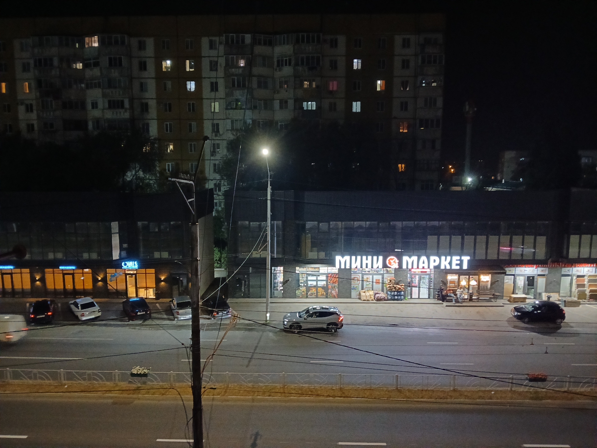 The image shows a nighttime street scene in Cherkessk, Russia, with a multi-story apartment building in the background and a row of storefronts in the middle ground. A street with several cars is in the foreground.
The apartment building is illuminated by numerous lit windows, suggesting occupancy. A street lamp is visible, casting light on the street below.
The storefronts include a "MINI MARKET" with illuminated signage and goods displayed outside, such as sacks and what appear to be pumpkins. Other storefronts are also visible, with illuminated entrances and windows.
Several cars are parked in front of the stores and on the street. One car, a silver SUV, is driving on the street. Another black car is parked in front of the MINI MARKET. There are also several parked cars to the left of the MINI MARKET, including a white SUV and a dark sedan. A white van is partially visible on the far left, moving away from the camera.
Two individuals are visible sitting on a bench outside the MINI MARKET. Another person is standing near a black car in front of the MINI MARKET.
The environment appears to be a typical urban street at night. Power lines are strung across the scene. A fence separates the sidewalk from the street in the foreground.