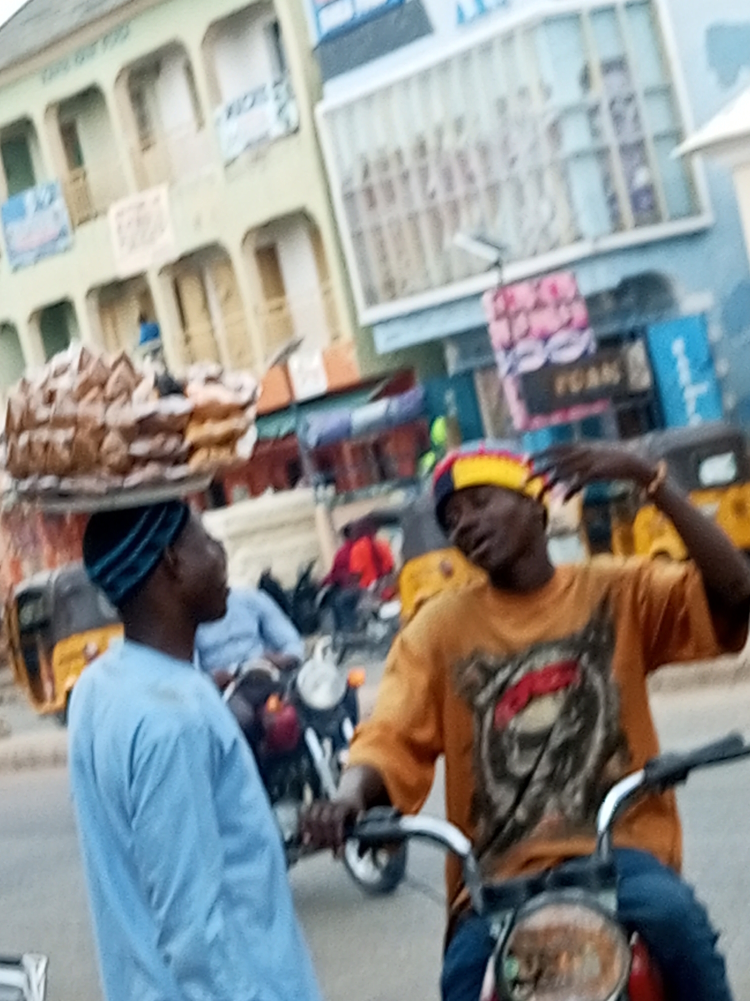 In Arawa, Nigeria, a street scene unfolds with a vendor balancing a large load of brown bags on their head, wearing a dark blue cap and a light blue traditional outfit. To their right, a young man on a motorcycle wears a colorful striped cap and an orange t-shirt featuring a graphic design. He is looking towards the camera with his right hand raised, palm facing outwards, in a gesture that could be interpreted as a greeting or a pose. The background reveals a street with various buildings and shops, including a building with arches and balconies and another with a large glass-fronted facade displaying posters. Several vehicles, including motorcycles and a three-wheeled auto-rickshaw, are visible on the street, suggesting a busy urban environment. The overall lighting suggests it is daytime.