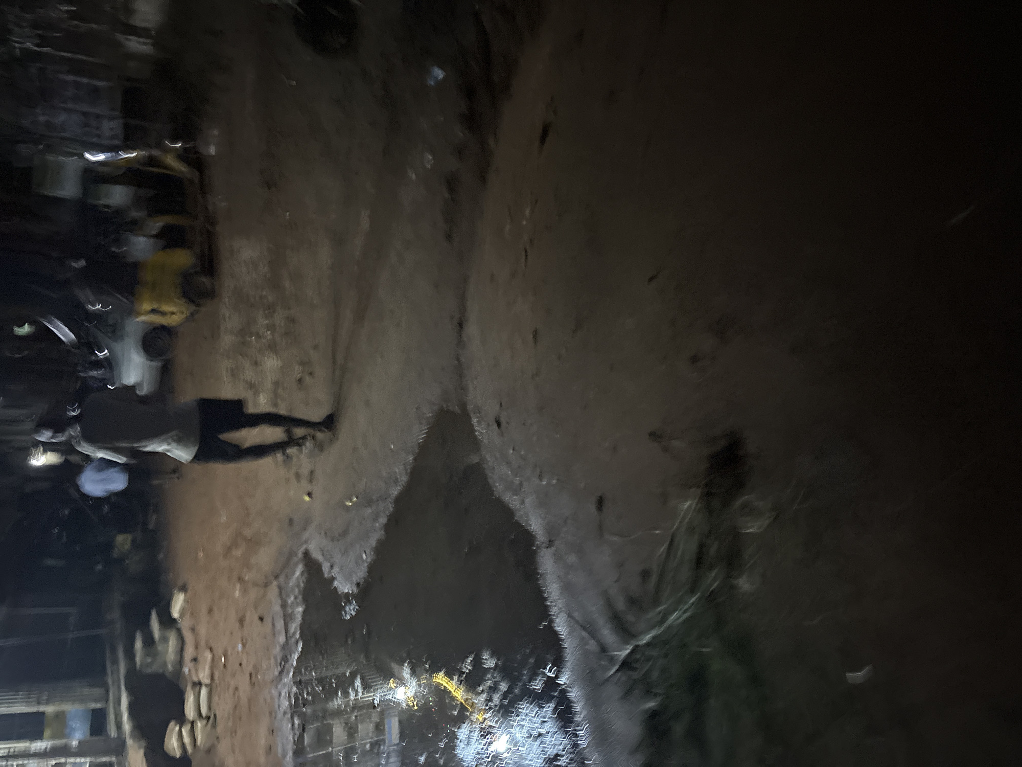 The image depicts a dimly lit scene at night, likely in a commercial or residential area of Onitsha, Nigeria. A person, silhouetted and facing away from the camera, walks on a dirt path. This individual is wearing dark shorts and a light-colored, short-sleeved shirt. Behind them, another figure is partially visible, wearing a light blue shirt.
The environment is characterized by a rough, unpaved ground with puddles of water reflecting faint yellow and white lights, suggesting artificial lighting from nearby structures or vehicles. Stacked sacks, possibly containing goods or construction materials, are visible to the left of the path. In the background, a yellow three-wheeled vehicle, common in Nigeria, is parked. The overall atmosphere is that of a bustling, yet somewhat unkempt, urban setting at night.