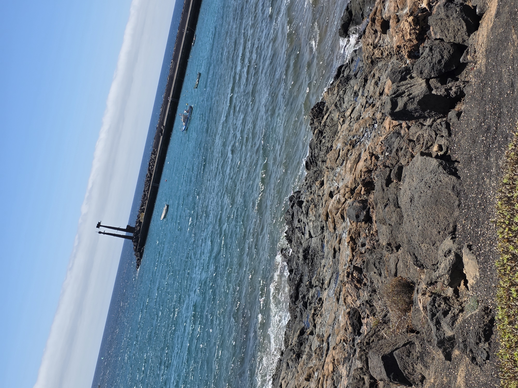 This image depicts a coastal scene in Teguise, Spain. In the background, a long, dark breakwater extends into the bright blue ocean, topped by two imposing, dark structures resembling pylons or masts. A few small boats are visible on the water near the breakwater, one of which is white and moored. The water is a vibrant turquoise with moderate waves. The foreground is dominated by a rocky, uneven shoreline composed of dark volcanic rocks and some reddish-brown stones. Sparse, dry vegetation is scattered amongst the rocks, and a line of low-lying green plants runs along the very bottom edge of the frame. The sky is a clear, pale blue with a long, thin band of white clouds stretching across it. The overall impression is of a sunny day at a rugged, natural coastline.