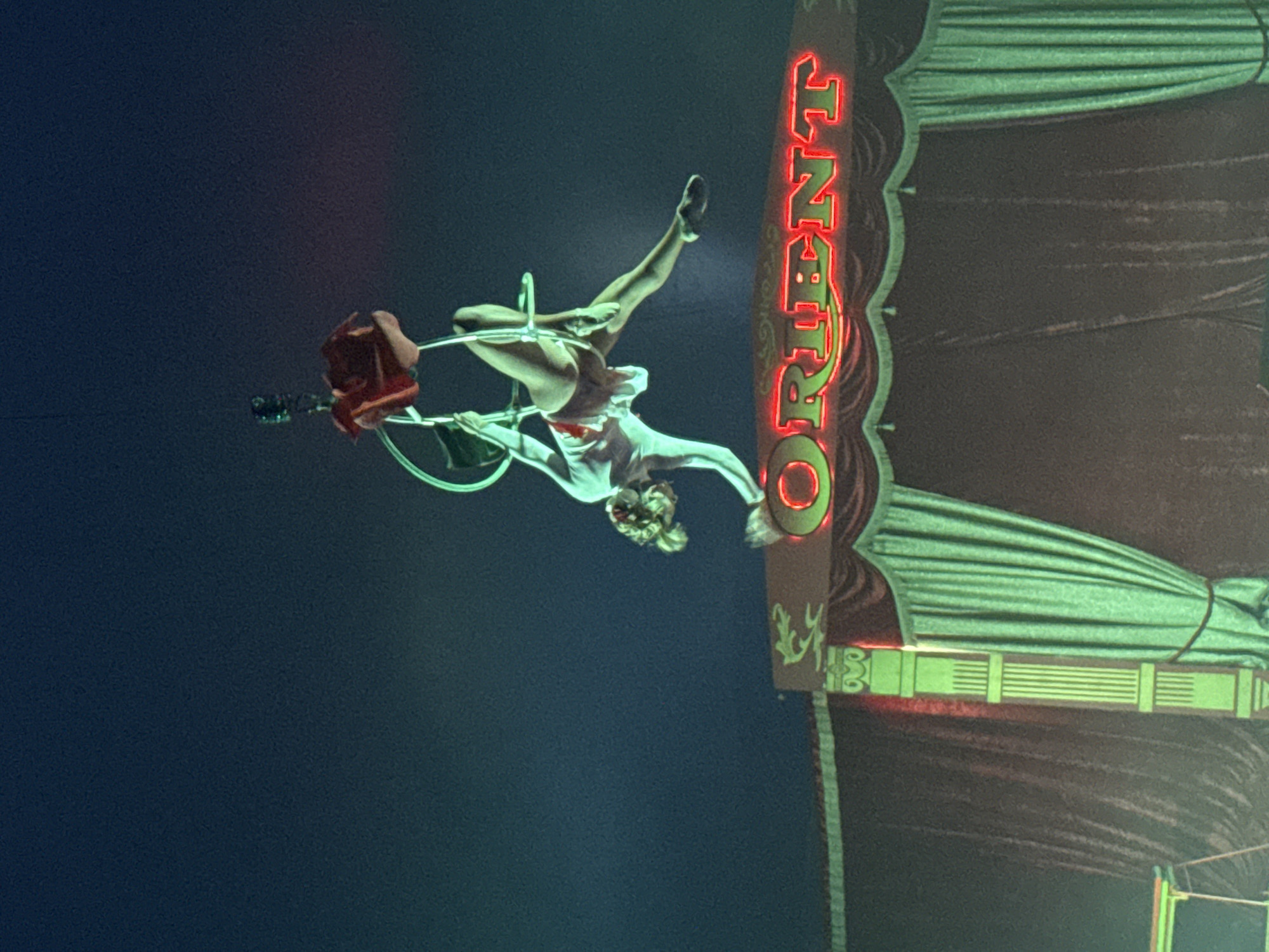 An aerial performer balances on a hoop suspended in the air at a show of the Orient Circus in Stara Zagora, Bulgaria. The performer wears a white top and short skirt, with one leg extended straight up in the air and the other bent, positioned inside the hoop. The word "ORIENT" in red neon lighting is visible in the background. The backdrop consists of green and maroon drapes.