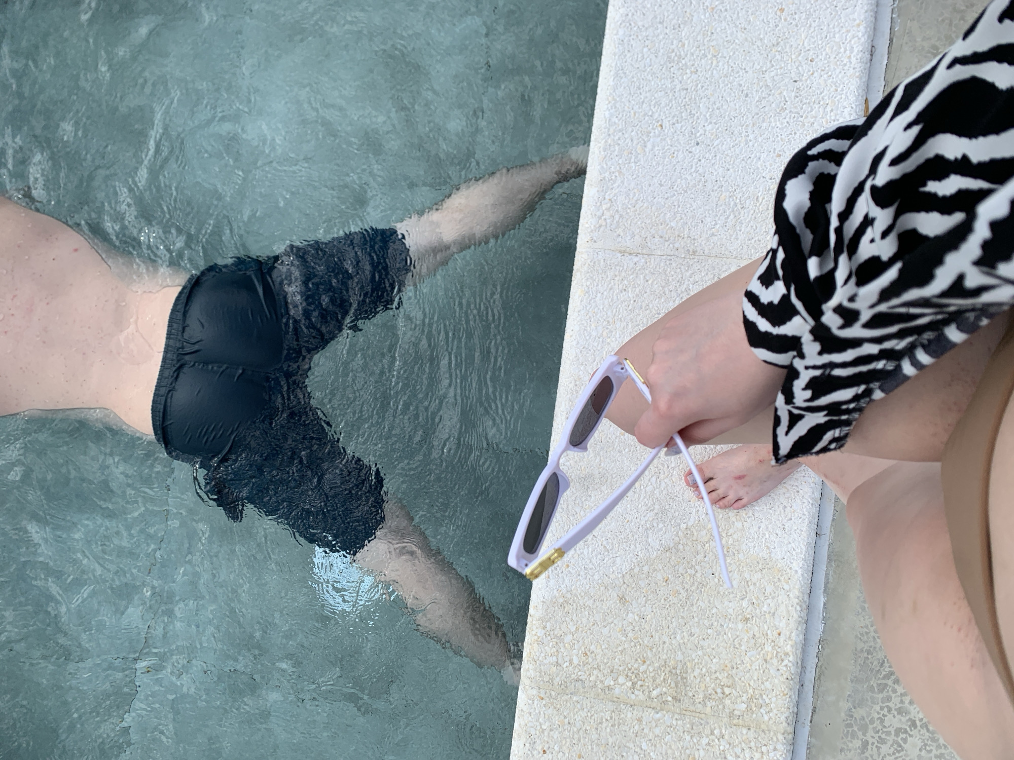 The image shows a person relaxing in a swimming pool. The scene is viewed from above, with the majority of the frame occupied by the pool water and the edge of the pool. A person's legs and torso are visible submerged in the water, wearing dark swim shorts. The water is clear and appears to be a pool with rippling surface.
On the edge of the pool, another person's legs and hand are visible. This person is wearing a black and white patterned garment, possibly a dress or top. They are holding a pair of white sunglasses with gold accents. The pool edge is made of textured white material.
The image appears to be taken during daylight, likely on a warm day given the swimming pool setting. There are no visible signs of specific weather conditions, but the lighting suggests it's not nighttime. The location cues are generic to a pool environment, with no specific landmarks or architectural features that would pinpoint a precise location within Severskaya, Russia. The overall mood conveyed is one of relaxation and leisure. No text is visible in the image.