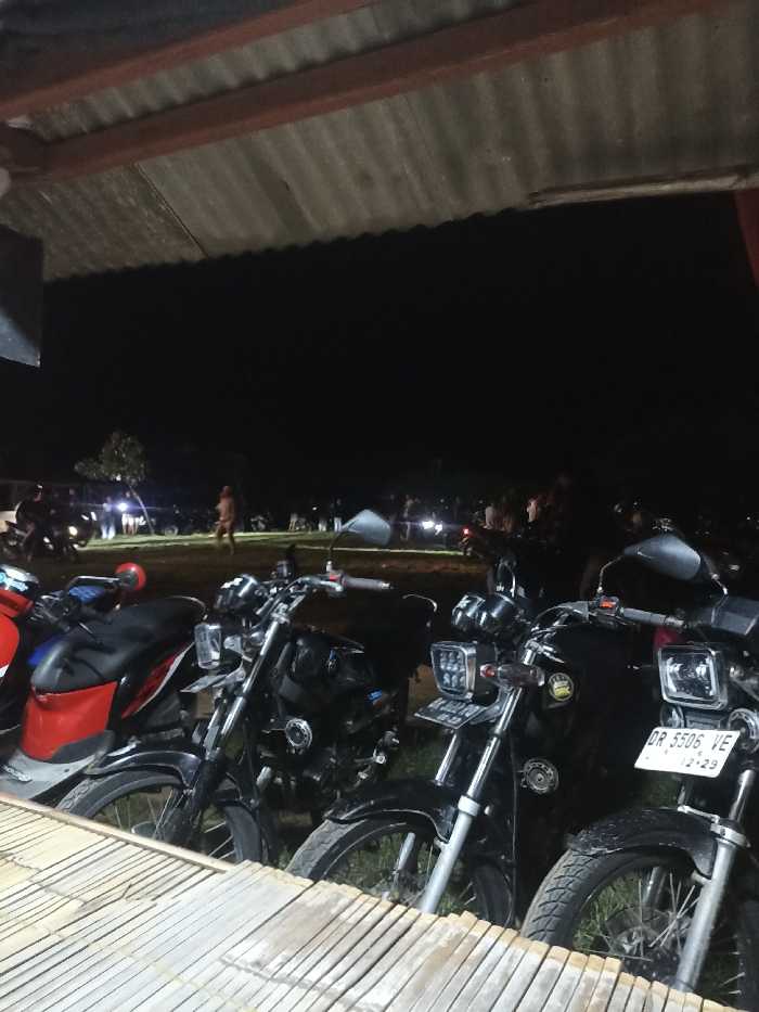 The image captures a nighttime gathering, viewed from beneath a corrugated metal roof. In the immediate foreground, a light-colored wooden or bamboo surface provides a base. Several motorcycles are parked in the mid-foreground, including a red and black scooter on the left, and at least three dark-colored standard motorcycles, some featuring square or rectangular LED headlights.
On the far right motorcycle, a white license plate reads "DR 5506 VE" with "12-23" below it, indicating an expiration date. A license plate on an adjacent motorcycle to its left partially shows "DL 4210". These license plate codes are characteristic of Indonesia, aligning with the provided location context of Kecamatan Pujut.
Beyond the parked motorcycles, an open dirt or grassy area is populated with numerous people standing and interacting in groups. One person stands somewhat centrally in the midground. Further in the distance, more figures and vehicle lights are visible against the dark night sky, suggesting a larger crowd or additional activities. The scene depicts a casual social event or meet-up, possibly for motorcycle enthusiasts, taking place outdoors or in a semi-sheltered space.