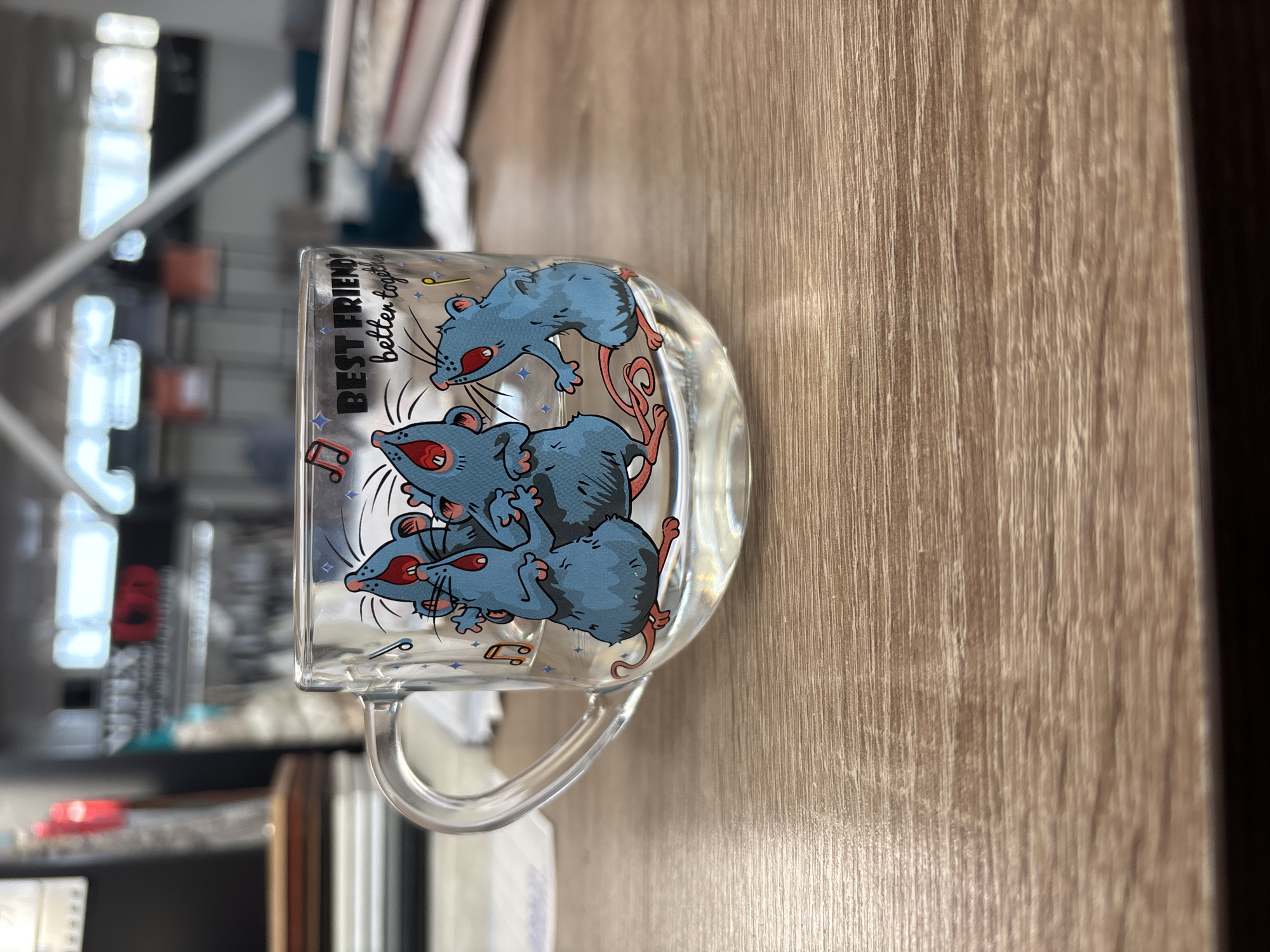 An indoor, medium-shot photograph taken at eye-level focuses on a clear glass mug on a wooden desk. The mug features a cartoon illustration of three blue mice singing with musical notes and stars around them. Above the mice, text reads "BEST FRIENDS better together." The background is blurred but suggests an office environment with shelves and lights. The lighting is bright, and the image is sharp, capturing the details of the mug and the texture of the desk.