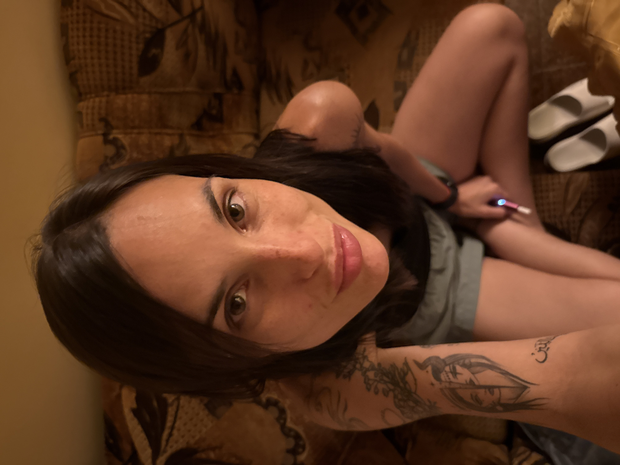 The image shows a woman with long, dark hair and light brown eyes, posing for a selfie. She has freckles on her face and a tattoo on her left arm. She is wearing a dark top and light-colored shorts. She is holding a pink and white object, possibly a pen or a small tool, in her right hand. The background appears to be a couch with a patterned fabric. The lighting is warm and intimate, suggesting an indoor setting. The location is not specified in the image or its context.
