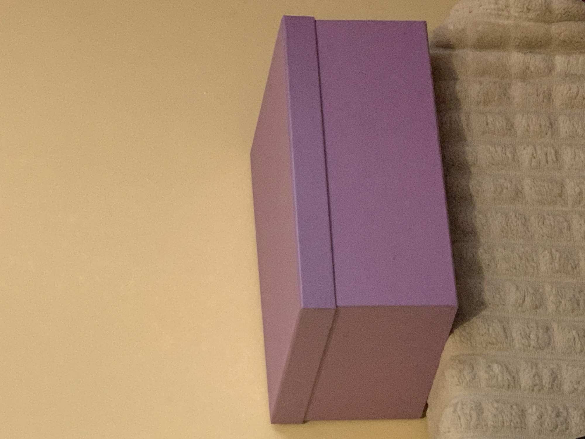 A close-up shot shows a rectangular purple box mounted on a wall. The wall is painted in a light tan color. To the right of the box is a light beige, textured material, possibly a towel or fabric with a repeating pattern. The photograph is taken indoors under artificial lighting.