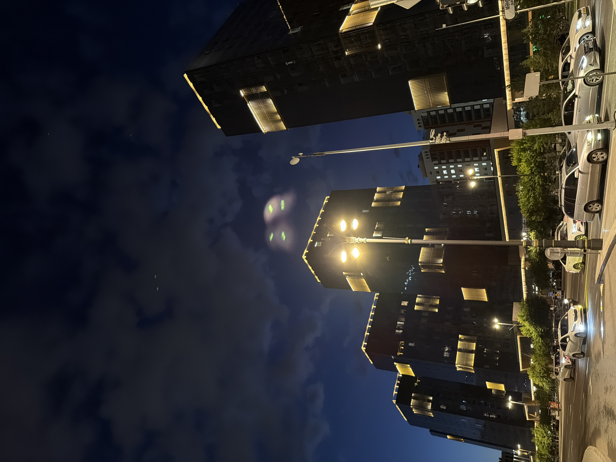 The photograph depicts a nighttime urban scene in Astana, Kazakhstan, characterized by modern architecture and street-level activity. Dominating the frame are several imposing, dark glass buildings with illuminated gold rectangular accents, suggesting contemporary office or residential structures. The sky above is a deep blue, transitioning to black, with scattered clouds and what appear to be two bright, circular lights, possibly aircraft or celestial bodies, visible in the upper portion of the image.
In the foreground, a multi-lane street is lined with vehicles, indicating traffic flow. Several cars are visible, including a white sedan with its headlights on, a silver station wagon, and other vehicles further down the road. The street itself appears wet, reflecting the lights from the surrounding environment, which may suggest recent rain. Streetlights, emitting a warm yellow glow, illuminate parts of the road and sidewalk. Lush green trees line the street, providing a natural element against the urban backdrop. The overall impression is one of a vibrant, modern city at night, with the architecture and lighting creating a striking visual effect.