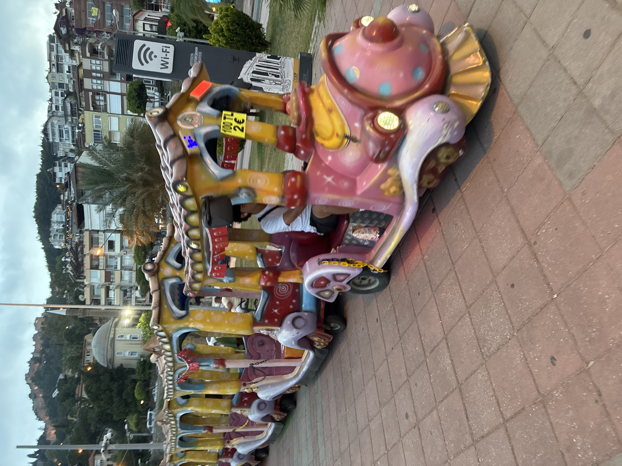This image displays a vibrant, multi-car children's trackless train ride set on a patterned reddish-brown paver walkway. The lead car of the ride is a whimsical, pink, polka-dotted teapot-like structure with golden accents and a spiraled tail, followed by several other uniquely designed, colorful carriages. A person, appearing to be a young adult, is seated in one of the middle cars, wearing a white t-shirt and black shorts.
A yellow sticker on the side of the ride indicates the cost: "100 TL" (Turkish Lira) and "2 €" (Euros).
In the background, a densely built-up cityscape sprawls across a green, tree-covered hillside, with numerous residential buildings climbing upwards under an overcast sky, suggesting it is late afternoon or early evening. A distinct domed building is visible amongst the distant structures. A tall black sign with a white "Wi-Fi" symbol is prominently featured, also displaying "ANTALYA BÜYÜKŞEHİR BELEDİYESİ" (Antalya Metropolitan Municipality), identifying the location as a public area within the Antalya region of Turkey. Palm trees are also visible among the buildings, adding to the coastal atmosphere. The scene captures a leisure activity in an urban, Mediterranean setting.