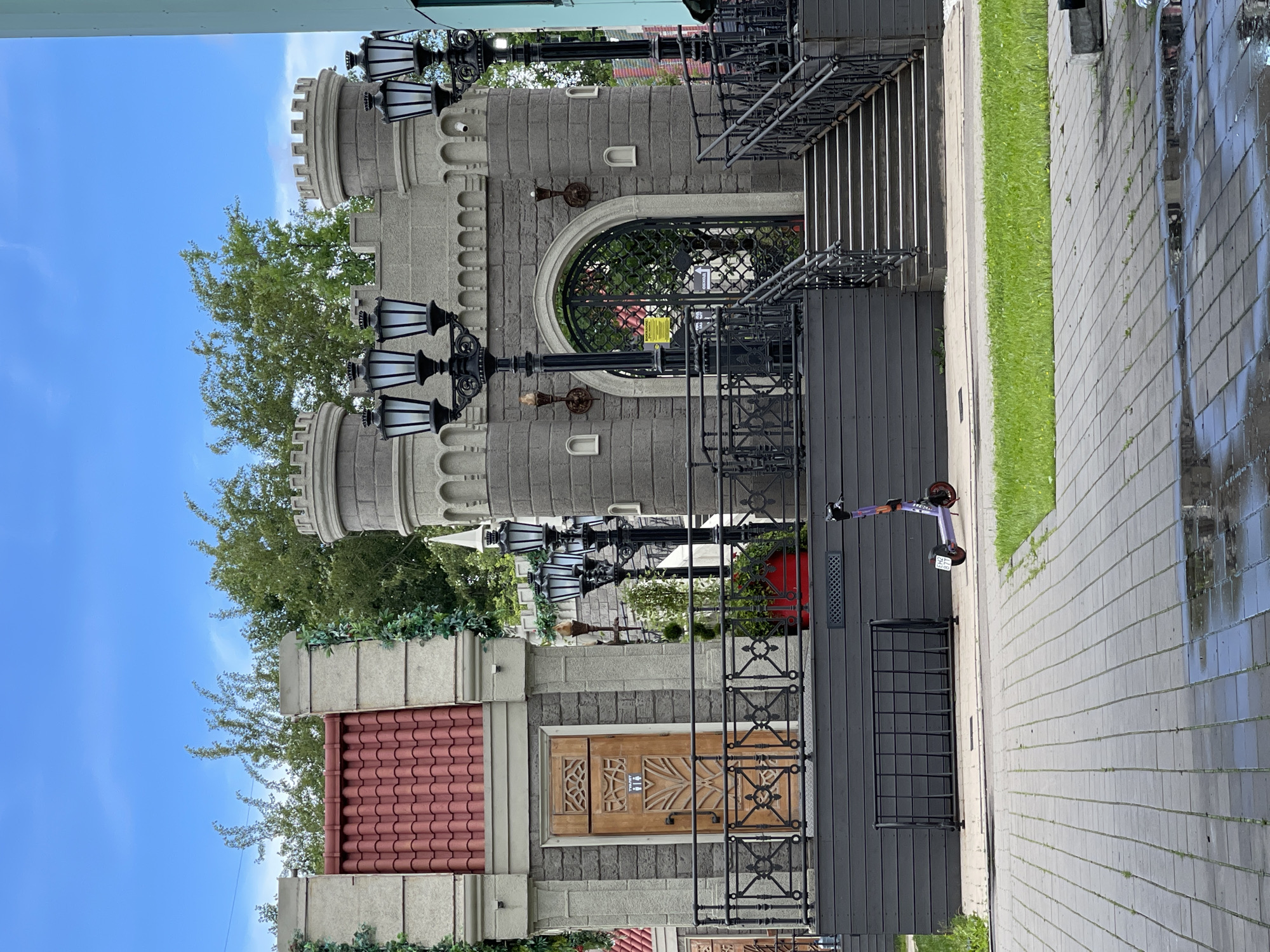 This image shows a castle-like building with stone walls and crenellated towers, reminiscent of a fairytale aesthetic. The building has decorative lamps and ornate metal railings. A wooden door with intricate carvings is visible. Above the door, a rounded window is covered with a black metal grate. Greenery, including trees and ivy, surrounds parts of the building. To the right of the building is a paved walkway with some wet patches, suggesting recent rain. A strip of green grass separates the walkway from the building. A purple electric scooter is parked near the building. The sky is blue with some clouds, indicating daytime. There is no visible activity taking place, and the scene appears peaceful and static. The building's architecture suggests it might be a restaurant, cafe, or a themed attraction. The overall impression is one of whimsical design in an urban or semi-urban setting. There is some text on the wooden door which appears to be an icon or symbol, possibly indicating a restroom. The image is taken from a slightly low angle.