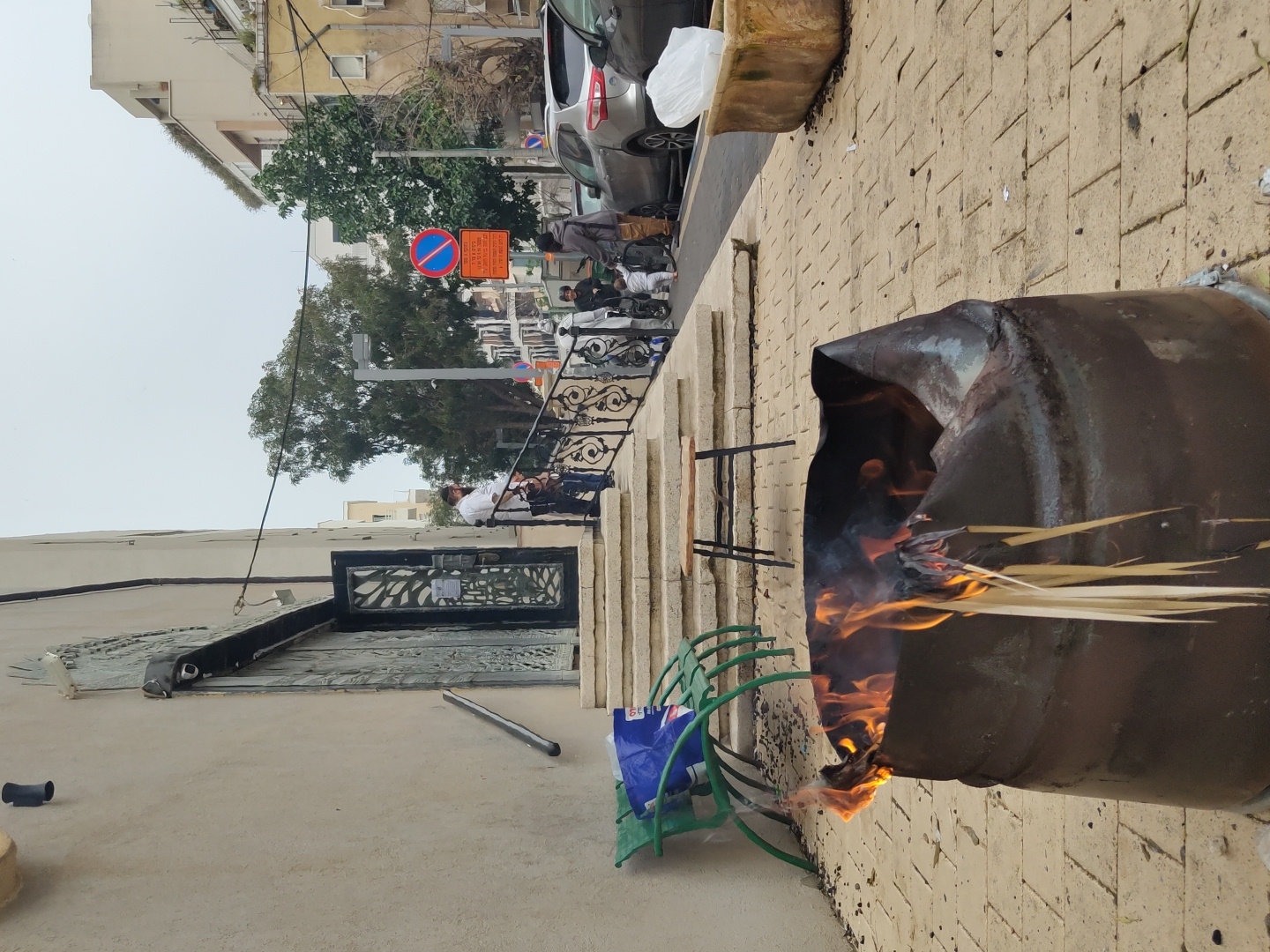 A partially destroyed metal barrel with flames is situated on a brick sidewalk, alongside a green chair and a blue plastic bag. In the background, a street scene includes pedestrians, cars, trees, and buildings. A "no parking" sign is visible. The location is Tel Aviv, Israel.