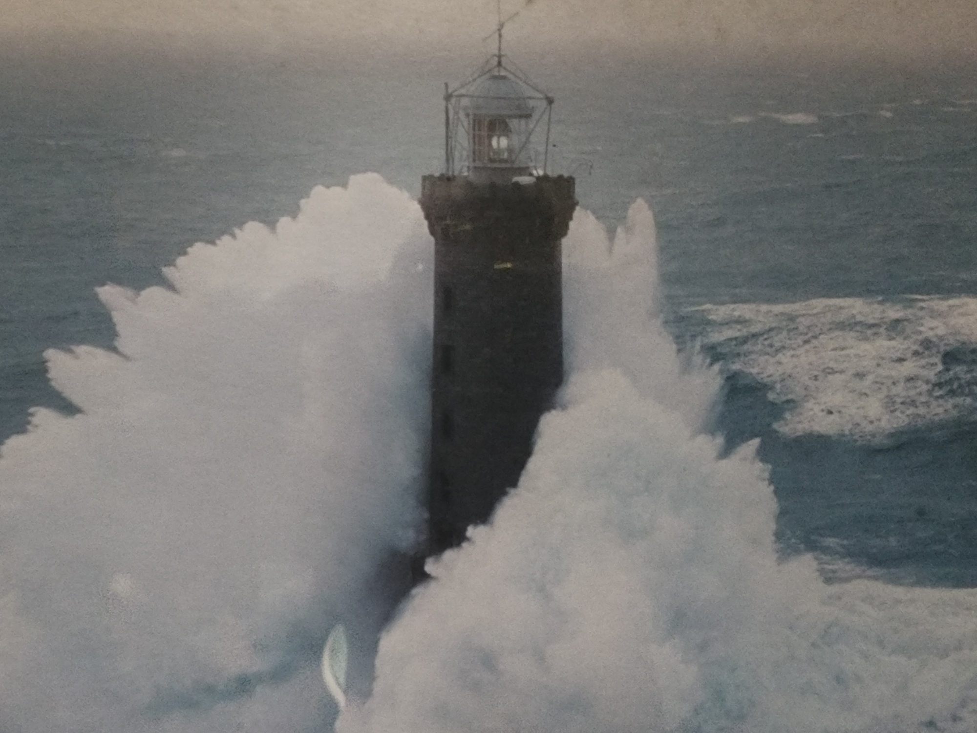 The image is a photograph of a lighthouse being hit by large waves. The lighthouse is tall and dark, with a light at the top. The waves are white and foamy, and they are crashing against the lighthouse with great force. The sky is overcast, and the sea is rough. The overall impression is one of power and drama. The photograph does not depict any specific location in Pyeongtaek-si, South Korea.