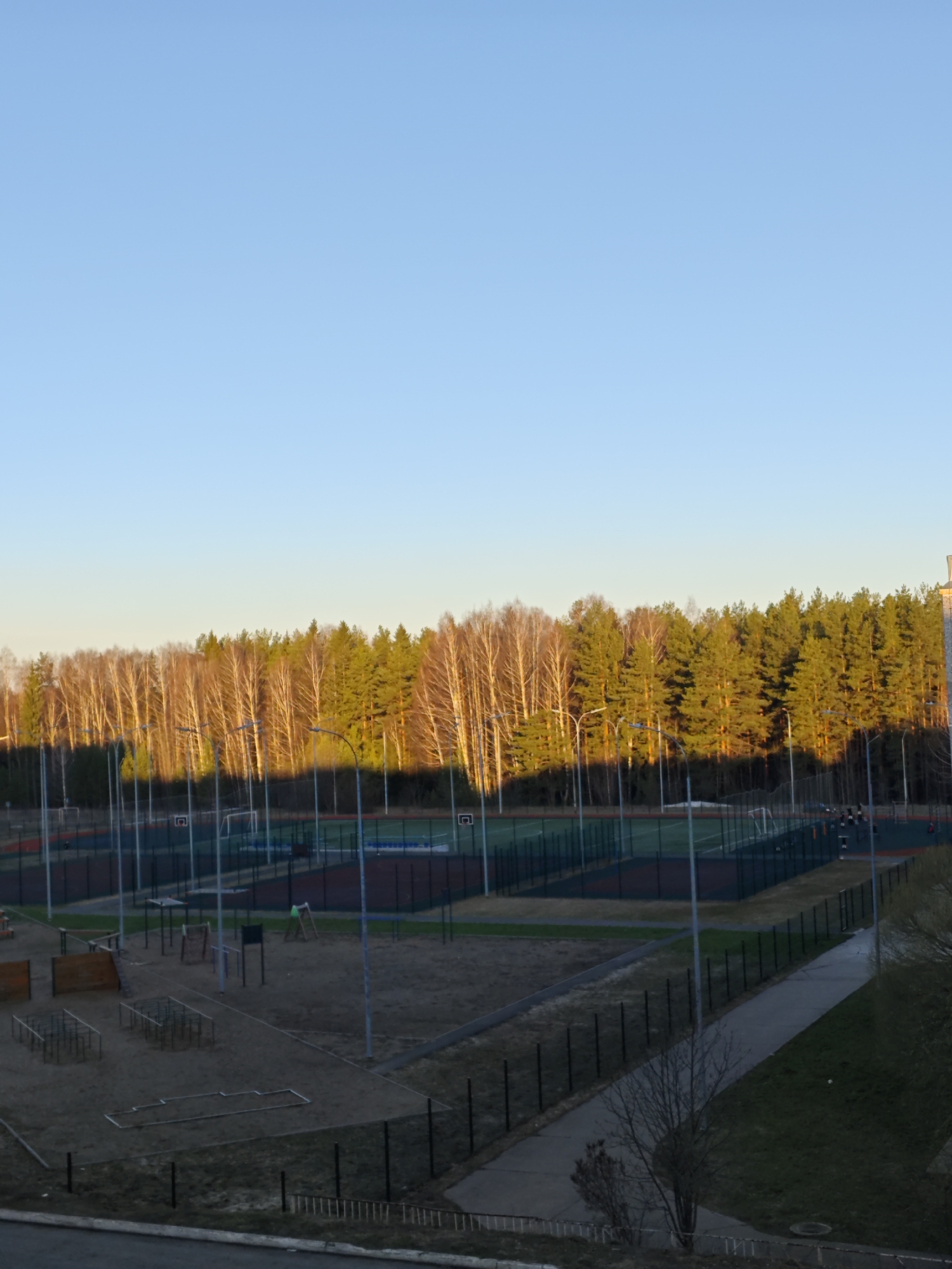 The image depicts an outdoor sports and recreation area in Markovskii, Russia. In the foreground is a sandy playground with metal climbing structures. Beyond this, several fenced-in sports courts are visible, possibly for basketball or volleyball. Tall metal light poles illuminate the area. In the background, a dense forest transitions from bare deciduous trees to evergreen pines, illuminated by the low-angle sunlight. A concrete walkway runs along the right side of the image. The sky is clear and blue.