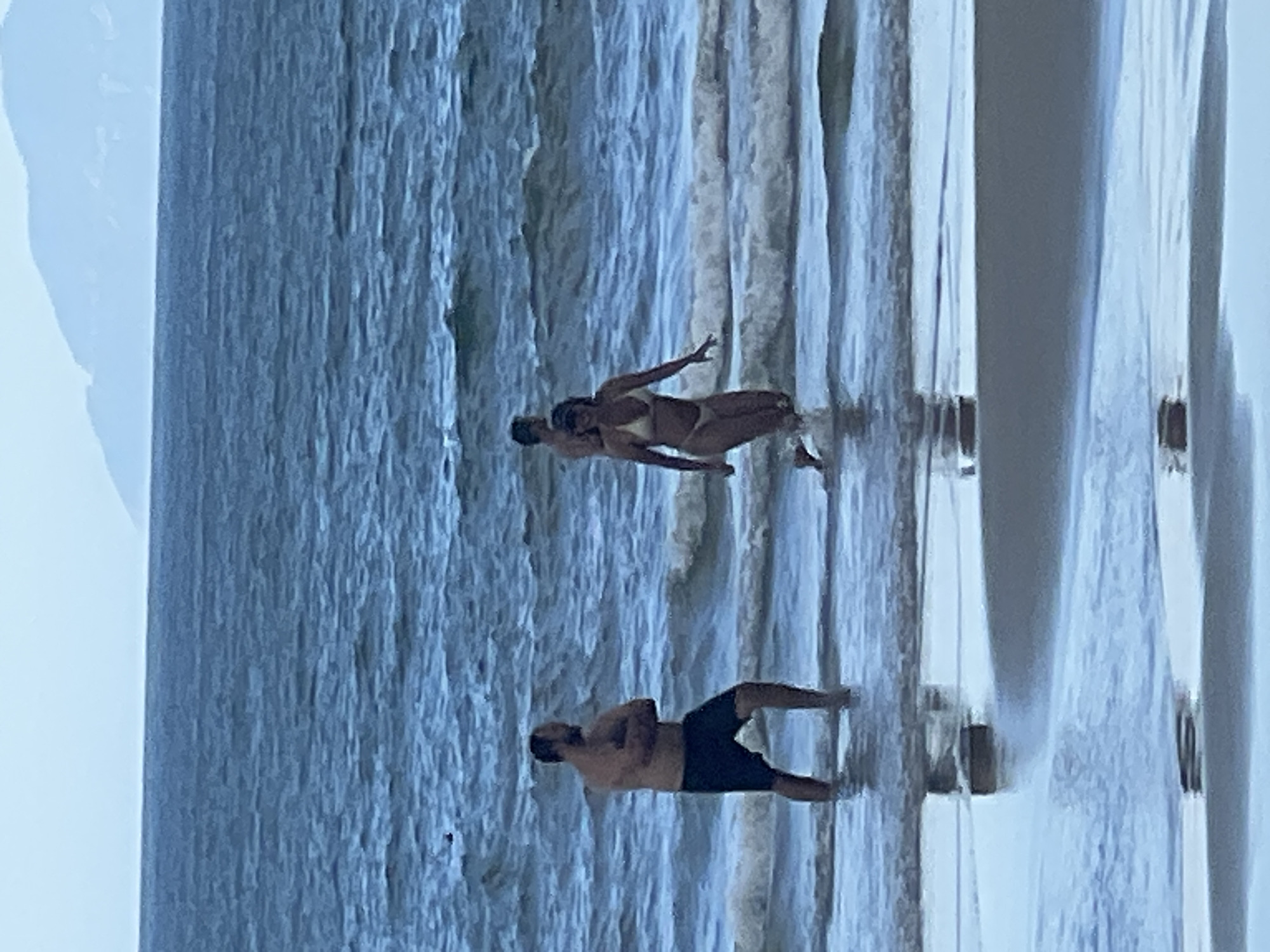 Three individuals are in shallow ocean water on a beach. A man in dark swim trunks stands with his arms crossed facing left. To his right, a woman in a light-colored bikini runs toward the viewer, her right leg extended. Behind her, a man with dark hair and a dark swim cap is visible in the water, partially obscured. The sea is blue with moderate waves. In the background, a hazy mountain range is visible under an overcast sky. The foreground shows a wet sand beach with shallow water reflecting the sky and sea. This scene is likely along the coast of Đà Nẵng, Vietnam.