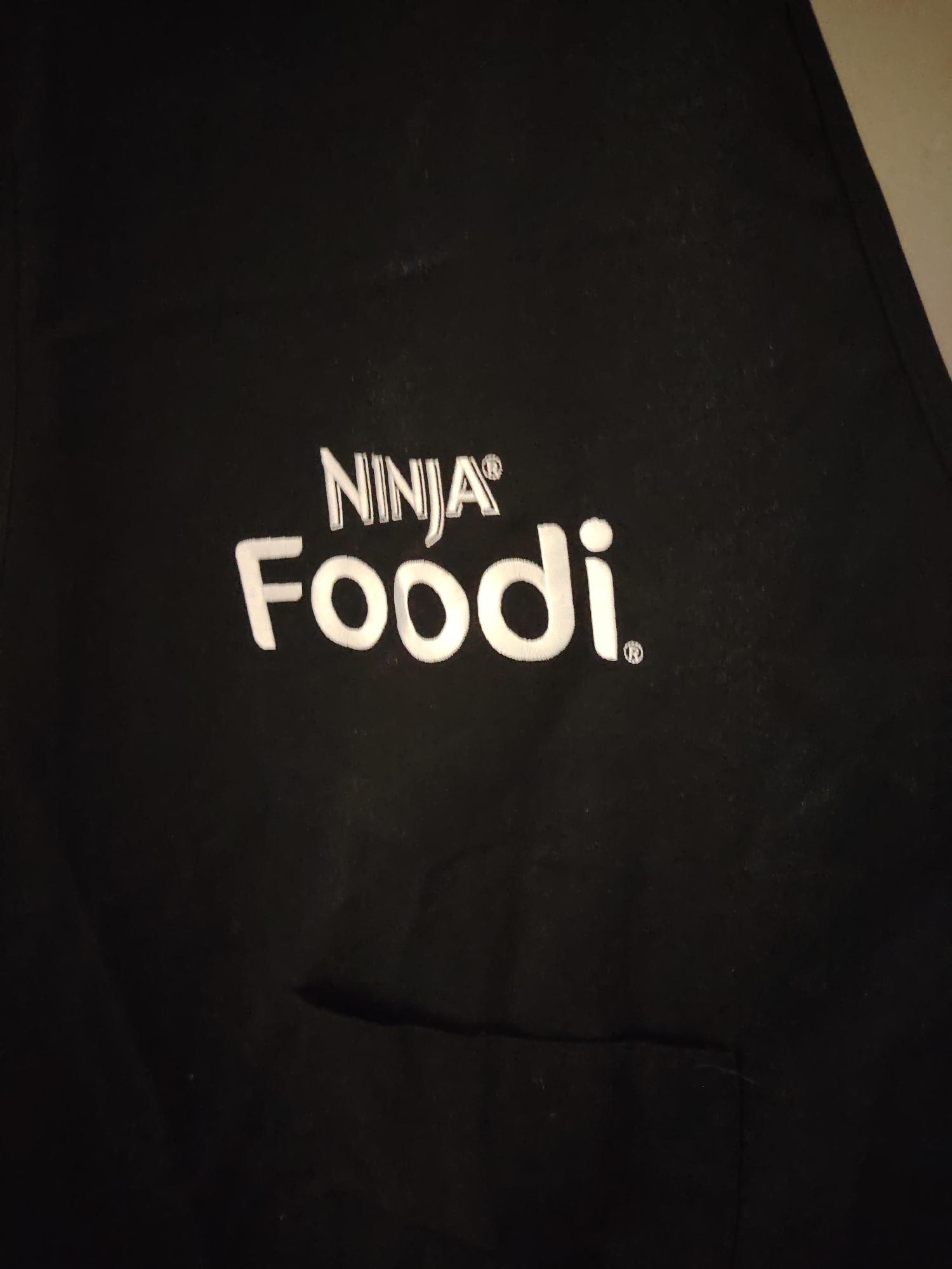 The image displays a close-up view of a black fabric garment, likely an apron, featuring white embroidered branding. The top line of text reads "NINJA®", positioned above the larger text "Foodi.®". Both brand names include the registered trademark symbol. A rectangular pocket is partially visible on the lower right of the garment. The fabric exhibits minor creasing and visible texture from the embroidery. No people or specific environmental details beyond the garment are discernible. This item is located in Huntingdonshire, United Kingdom.