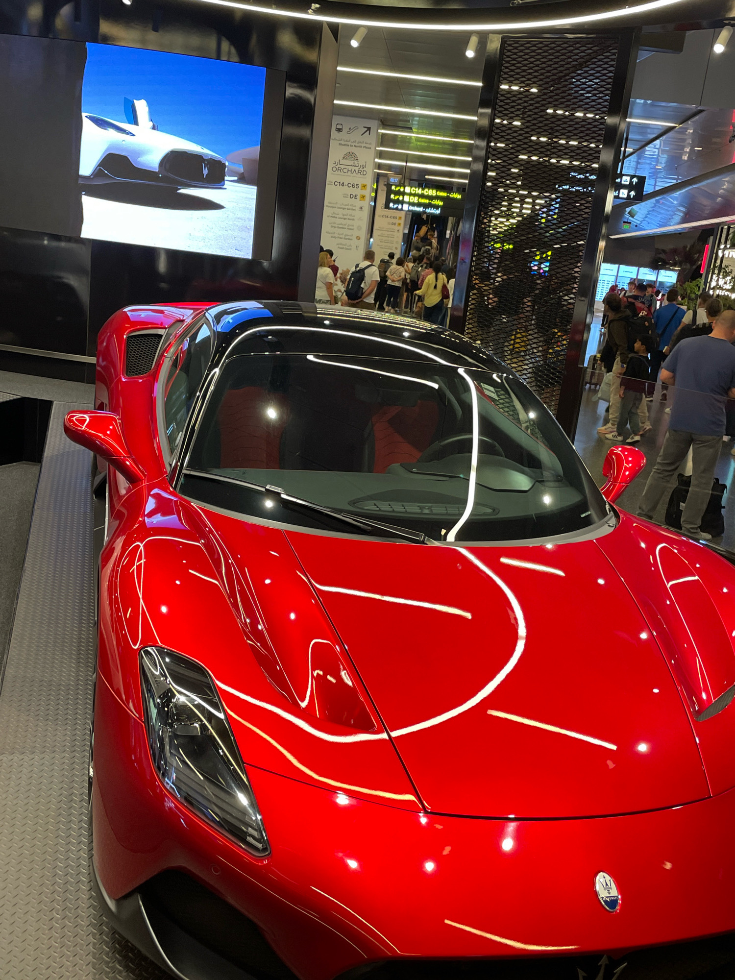 A bright red sports car, possibly a Maserati, is displayed on a platform in a modern indoor environment. The car is positioned diagonally, with its front left corner closest to the viewer. The car's hood is highly reflective, showing streaks of white light from the overhead lighting. The interior appears to be a public space with many people walking around. In the background, there's a large screen displaying an image of a white car, and a series of signs, some in Arabic and English, indicating directions. One sign reads "ORCHARD" and lists gate numbers like "C14-C65". This scene is located within the Hamad International Airport in Doha, Qatar, a major international hub known for its modern architecture and luxury amenities.