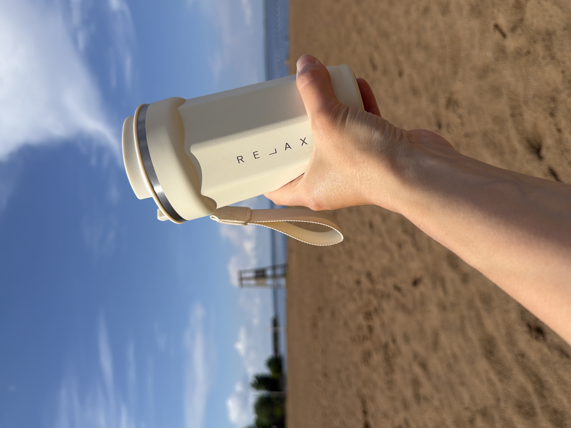 A person's hand is holding a cream-colored tumbler with the word "RELAX" printed vertically on its side. The tumbler has a strap attached to its lid. The background is a sandy beach with a body of water and a lifeguard tower in the distance. The sky is blue with some clouds. The image appears to be taken in Chaikovsky, Russia, given the presence of a lifeguard tower which is common at the beaches along the Kama River in that city.