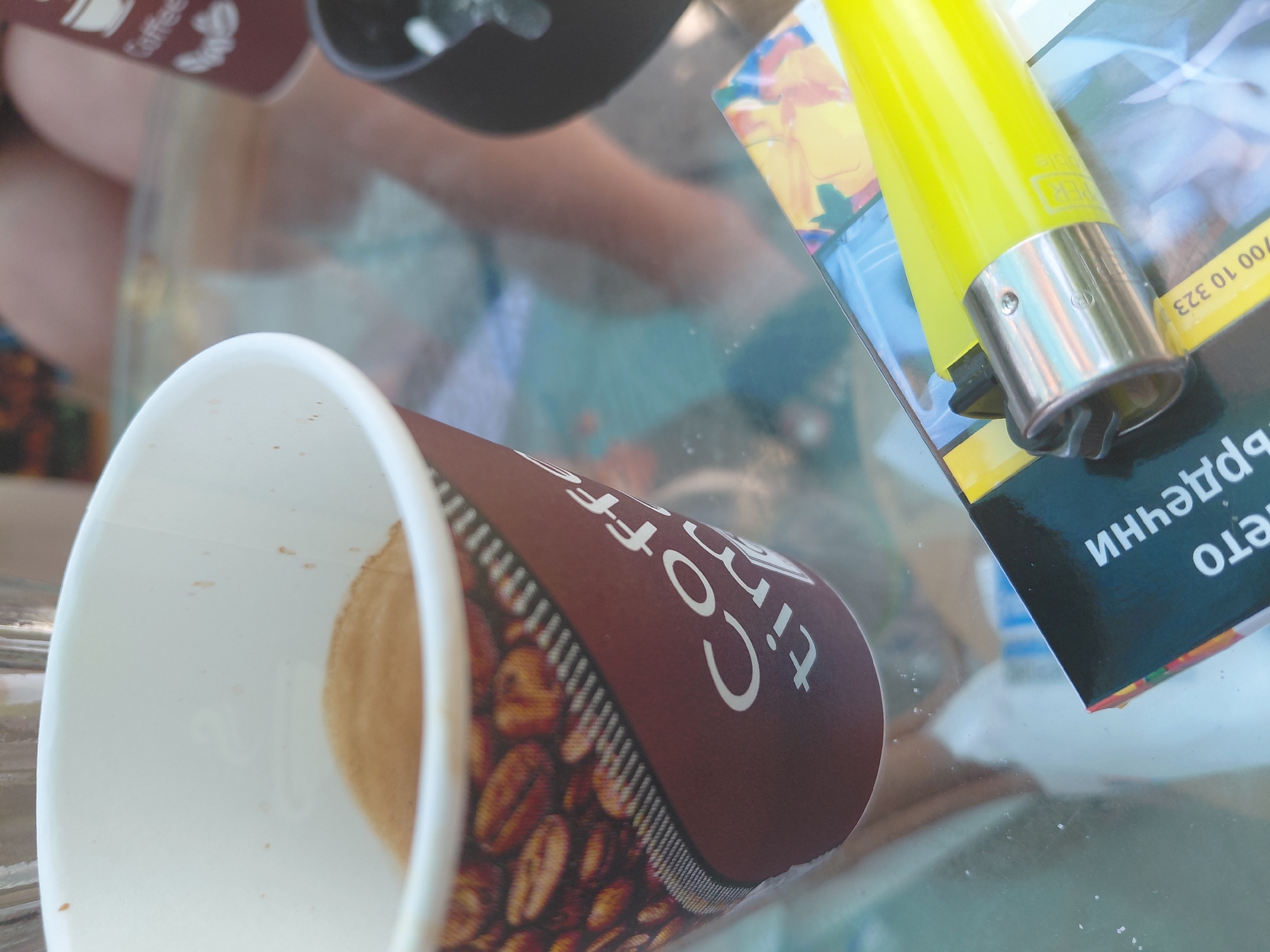 The image presents a close-up, high-angle view of a tabletop scene in Kostievo, Bulgaria. A disposable coffee cup, white on the interior and brown on the exterior with coffee bean graphics and partial "Coffee time" text, contains a small quantity of light brown residual liquid or crema. Resting adjacent to the coffee cup, on a black cigarette pack with yellow and red accents, is a bright yellow Clipper brand lighter featuring a metallic top and a black flint wheel. The cigarette pack displays Cyrillic text, including part of a warning label "ИННЕЧЬДР" and numbers "00 10 323," consistent with Bulgarian packaging. The objects are situated on a clear glass table, which reflects blurred imagery of the environment and the lower extremities (legs/feet) of at least one individual. In the out-of-focus background, another brown disposable coffee cup and a dark, cylindrical object are partially visible. The scene suggests a casual outdoor or semi-outdoor setting.