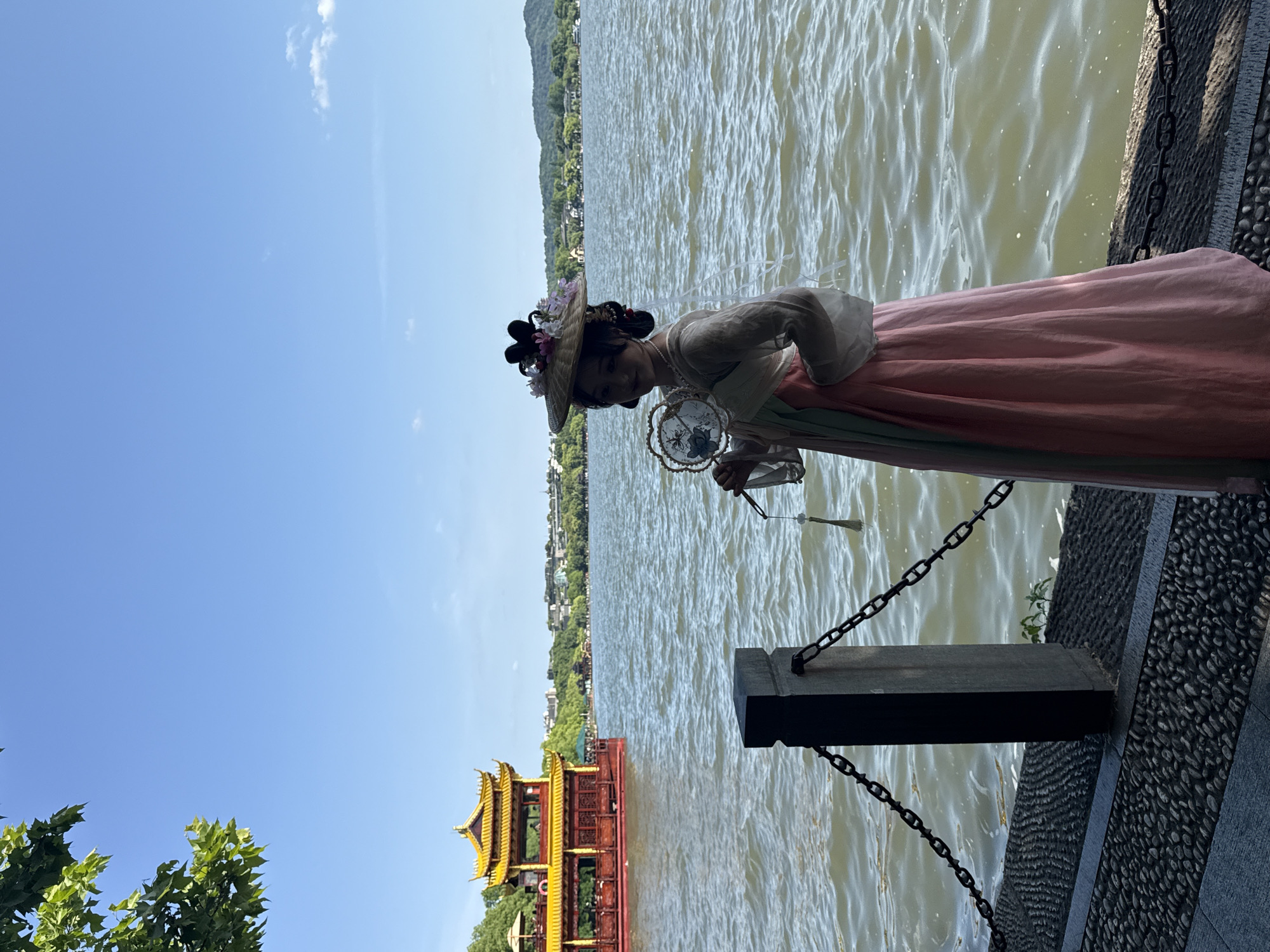 A woman in traditional Chinese clothing stands by the lake in Shangcheng District, China, holding a decorative fan. She is wearing a pink dress with green accents and a hat adorned with flowers. In the background is a body of water with a building on a boat, and trees line the shore. The sky is blue with few clouds. A chain-link fence borders the water.