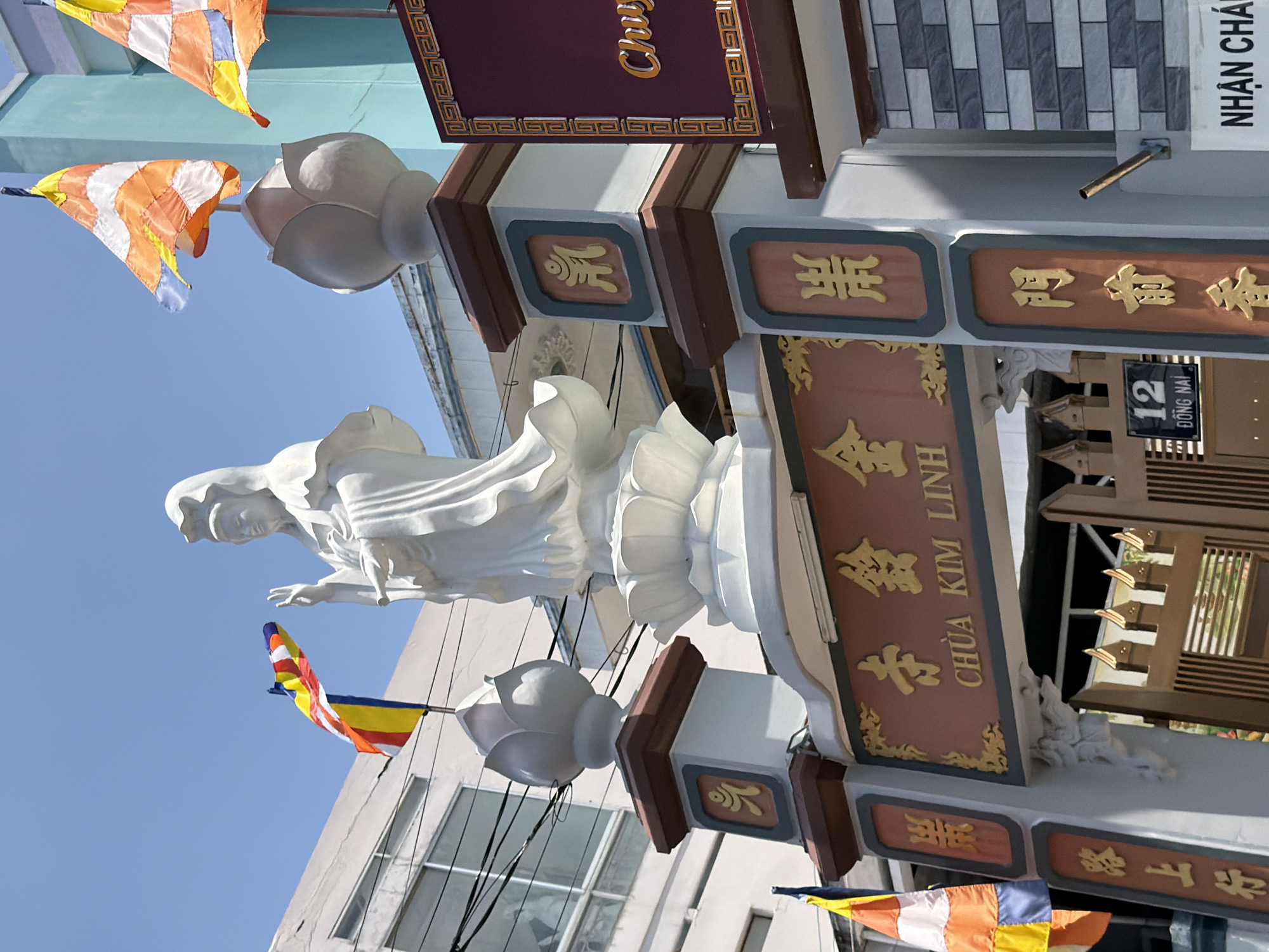 A white, serene statue of a female deity stands prominently in front of a bright blue sky. The statue is adorned with traditional Buddhist prayer flags in various colors, including orange, yellow, blue, and white, fluttering in the wind. One flag is positioned near the deity's head, while another is visible to the left. The statue is situated on a pedestal that is decorated with lotus motifs.
In the foreground, a gate, partially visible, features ornate carvings and traditional Vietnamese calligraphy. The gate has a brown wooden structure with decorative metal elements. Above the gate, a sign with gold lettering in Vietnamese reads "CHUA KIM LINH." To the right of the gate, a building with blue and light blue colored walls can be seen. The environment appears to be a temple or religious site, possibly in Vietnam, given the script and prayer flags. The presence of electrical wires suggests an urban or semi-urban setting. A street sign with "12 DONG NAI" is also visible near the gate. The overall scene is brightly lit, suggesting it is daytime.