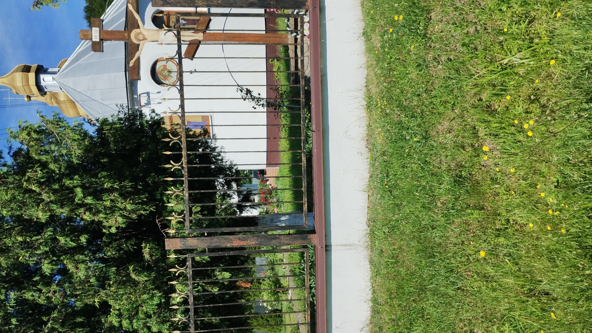 The image shows a portion of a churchyard in Chynadiiovo, Ukraine. In the foreground, a grassy area with scattered yellow wildflowers leads to a white, low wall topped with a dark brown decorative border. Behind this wall is a black wrought-iron fence with ornate golden accents on its posts and the tops of the vertical bars. Beyond the fence, a white church building with a silver roof and golden domes is visible. A wooden cross with a crucifix of Jesus Christ is positioned in front of the church building. To the left of the church, a large green tree provides shade. The sky is a clear blue. The scene appears to be peaceful and well-maintained.