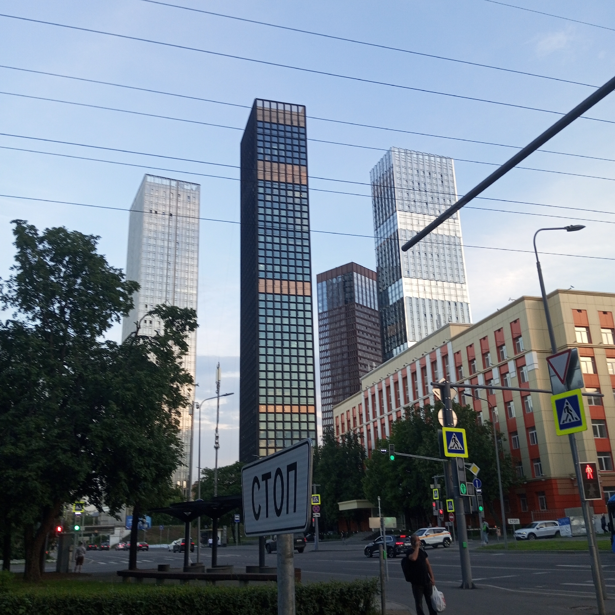 The image captures a street scene in Moscow, Russia, likely during the daytime with a clear sky. Dominating the background are several modern skyscrapers, some with glass facades reflecting the light, and one with a distinct pattern of darker and lighter panels. In the foreground, a street sign with the Cyrillic word "СТОП" (STOP) is prominently displayed on a metal pole. Traffic lights are visible, with one showing a red signal and another a green signal. A pedestrian crossing sign with a blue background and a white silhouette of a person is also present. Several cars are on the road, indicating active traffic. A man is walking on the sidewalk, carrying a plastic bag. The environment includes trees, a building with red and beige accents, and overhead power lines.