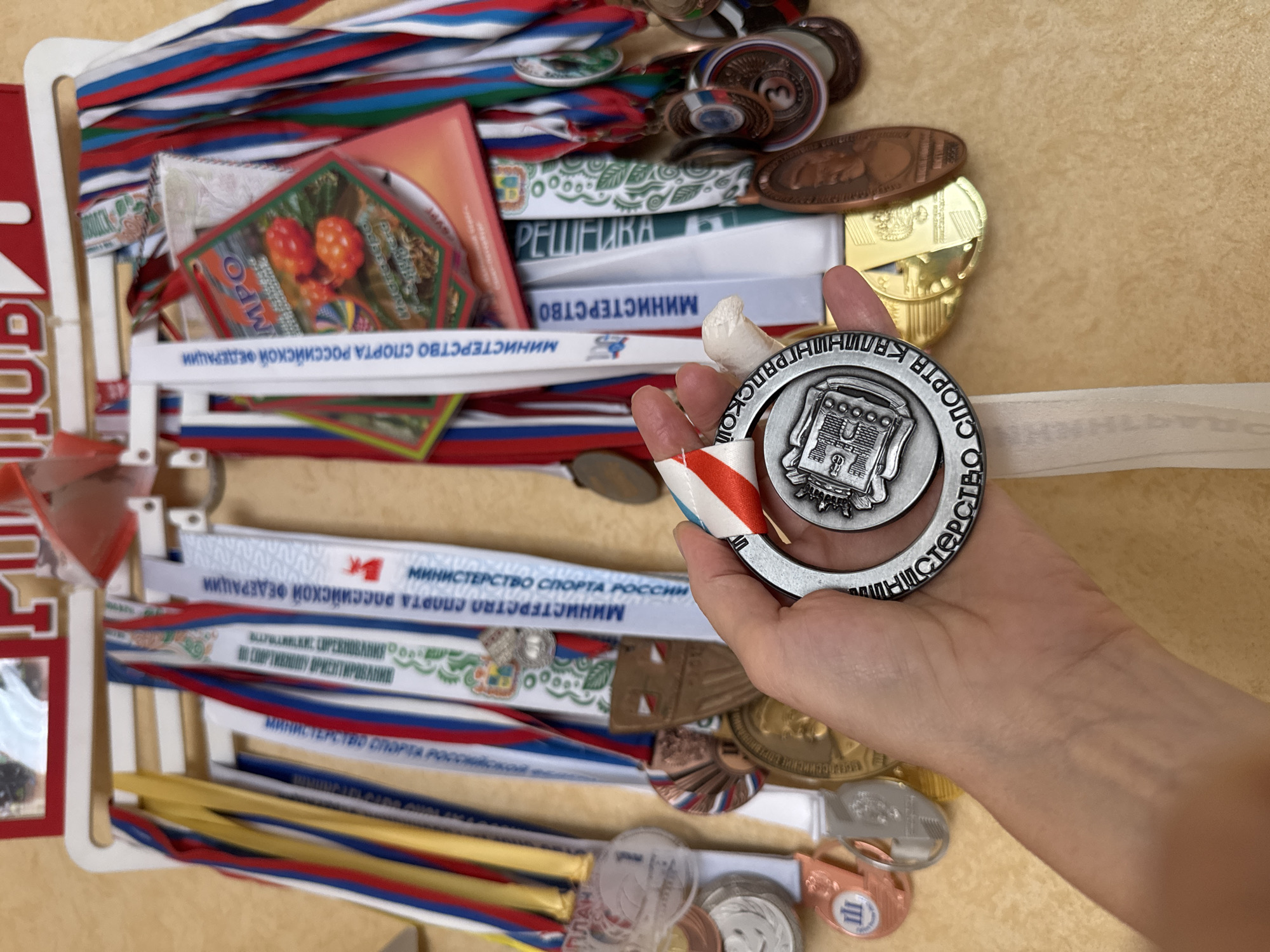 Here is a detailed description of the image:
**Overview:**
The image shows a collection of medals and awards, some of which are held in a person's hand. The medals appear to be related to sports achievements, and the scene suggests a celebration or recognition of athletic accomplishments.
**Details:**
* **Hand:** A person's hand is holding a silver medal with what seems to be the image of an architectural structure. There is a white and red-and-blue ribbon attached to the medal. The finger is partially wrapped in tape.
* **Medals and Awards:** There is a collection of medals and award ribbons on a white medal holder.
* **Russian Flags:** The Russian flag is used to design most of the medal ribbons.
* **Text in Russian:** Text in Russian is visible on many of the medals and ribbons, indicating that the awards are likely from Russia. The text includes phrases such as "Ministry of Sport of the Russian Federation."
**Overall Impression:**
The image is a display of sports achievements, likely from Russia, with a focus on the medals and recognition received by athletes.