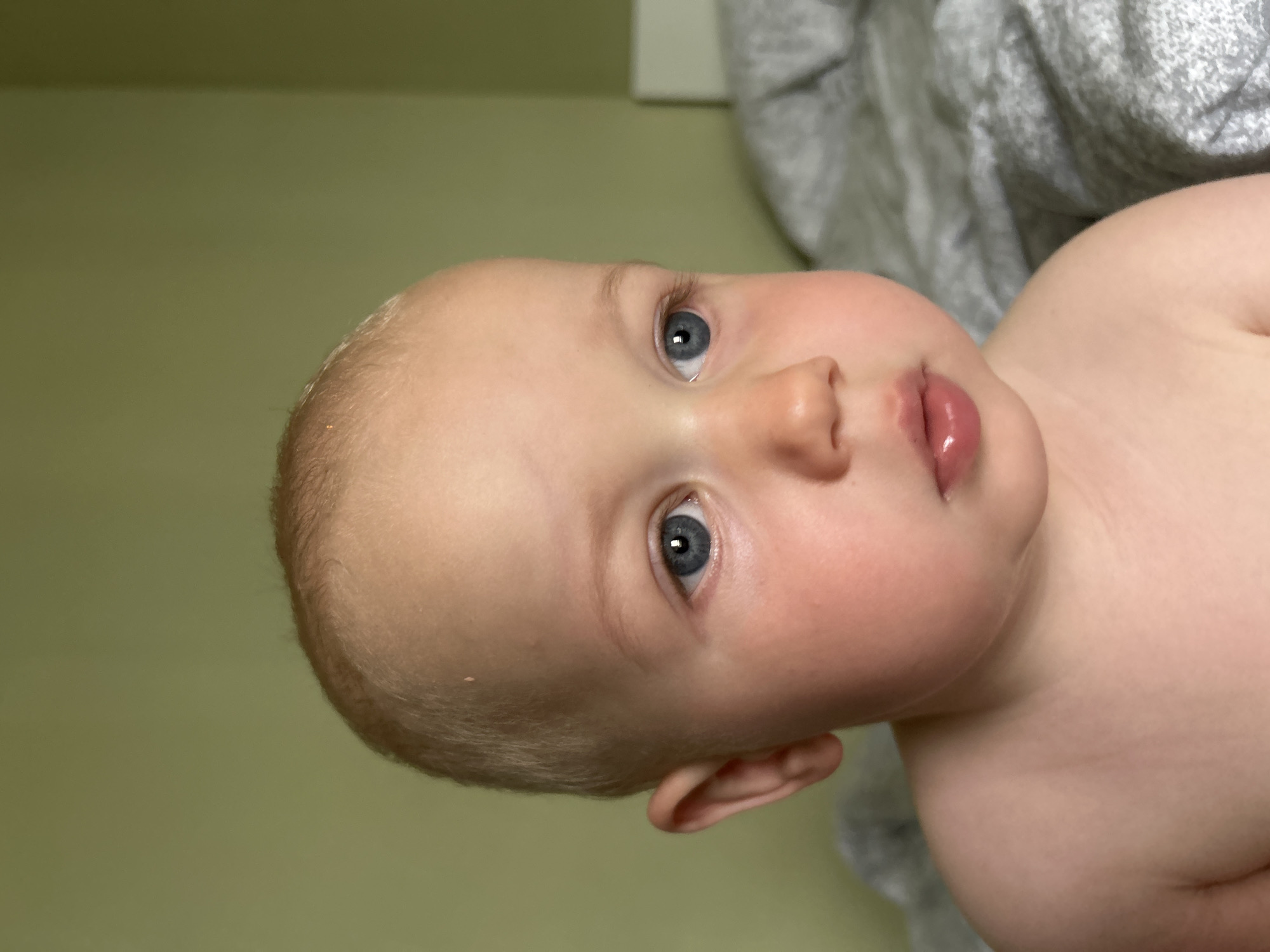 A close-up, medium shot shows the head and upper body of a fair-skinned infant with fine, light blonde hair. The infant's face is turned to the right, with blue eyes looking to the right. Rosy cheeks are visible, as are small, closed lips. The infant's bare shoulder and upper chest are visible in the lower portion of the image. The background consists of a solid pale green wall, and a grey, textured surface, likely bedding, on the right side. The image is a photograph taken indoors. No specific location in the city of Kaluga, Russia can be determined from the visual information provided.
