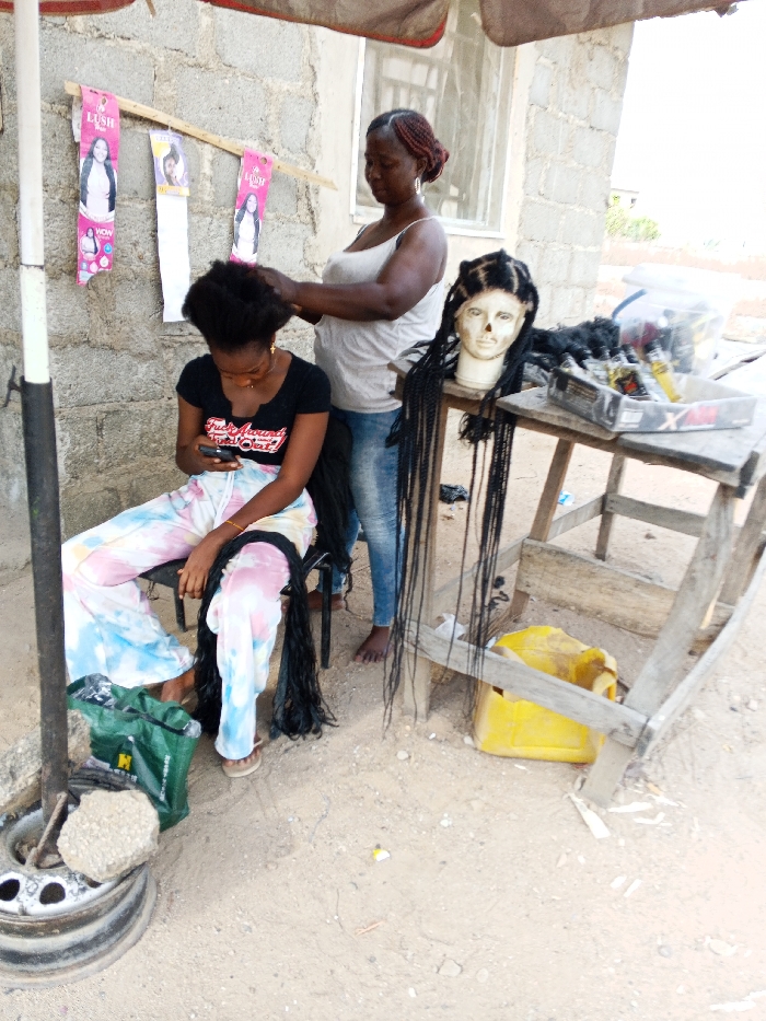 The image depicts an outdoor hair braiding operation in Adonkolo, Nigeria. An adult female, barefoot and dressed in a white sleeveless top and blue jeans, stands behind a seated younger female. The adult female is actively braiding the younger female's dark, voluminous hair. The younger female, wearing a black t-shirt with text, tie-dye sweatpants, and light-colored flip-flops, is focused on a smartphone held in her hands. Strands of long, dark braided hair extensions are visible on her lap and extending to the ground.
To the left, a dark metal pole, supported by a car tire rim filled with concrete, holds a dark-colored overhead tarp for shade. Three packages labeled "Lush" are affixed to a horizontal wooden stick against a grey cinder block wall. To the right, a light-colored mannequin head featuring long, dark braided hair extensions rests on a rustic wooden table. A black tray containing hair products and tools is also on the table. A yellow plastic jerrycan is positioned on the ground beneath the table. A green plastic bag is visible on the sandy ground near the seated client's feet. The background consists of a cinder block building with a window, and sparse vegetation is visible in the distance to the upper right. The ground is composed of light brown sand and dirt.