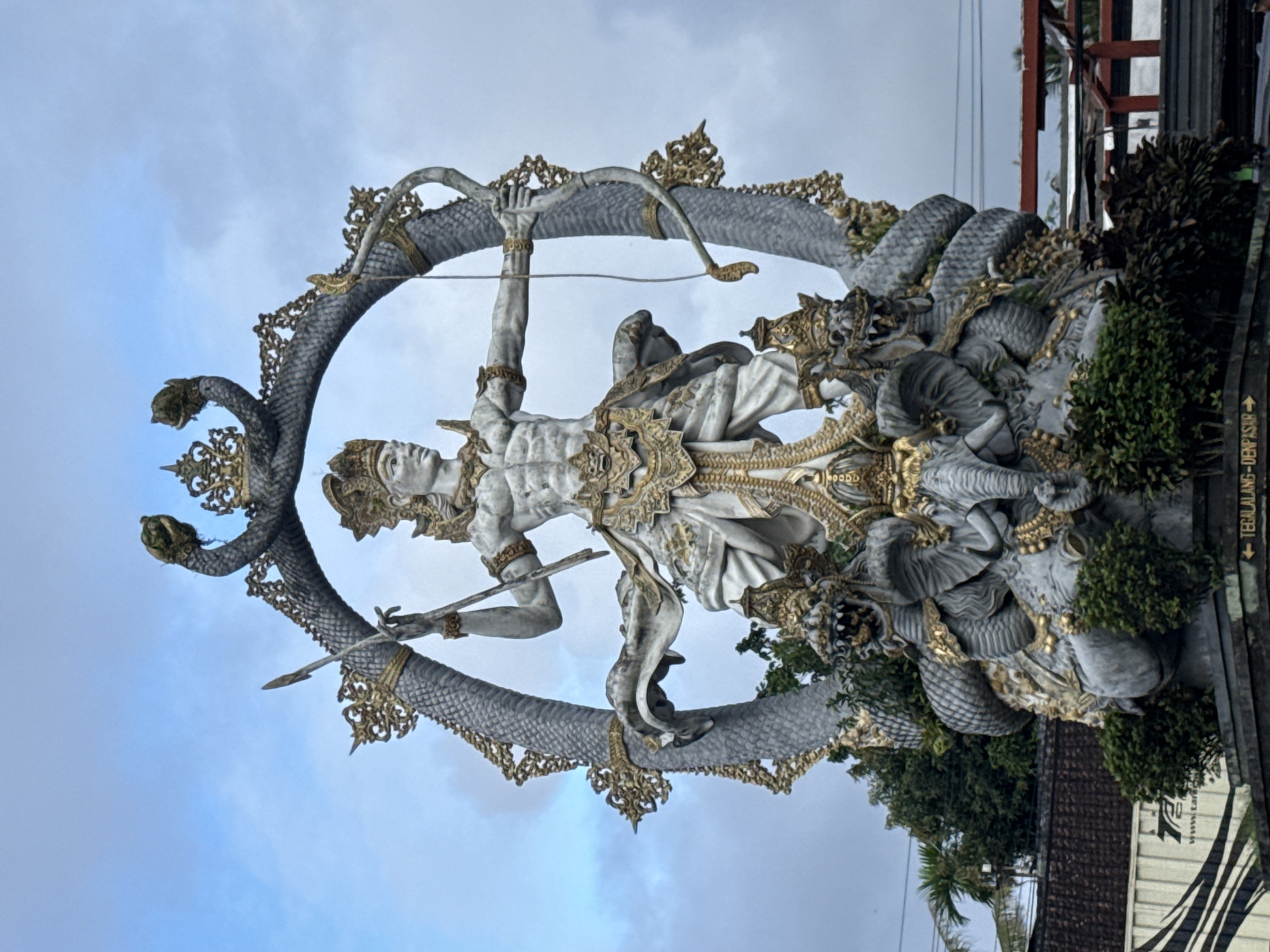 The image shows a large, ornate sculpture of Arjuna, a hero from Hindu mythology, located in the Ubud District of Indonesia. Arjuna is depicted as a pale, muscular figure adorned with golden jewelry and a crown. He holds a bow in his left hand, raised above his head, and a sword or dagger in his right hand, angled downward. The sculpture is encircled by a large, gray serpent with multiple heads, its scales intricately detailed and accented with gold ornamentation. The serpent forms a circular frame around Arjuna, with its tail and head positioned on either side of the figure. The sculpture is partially obscured by green foliage and a dark-colored structure in the background. The sky is a pale blue with wisps of clouds. A directional sign with some inscriptions is visible on the right side of the image.