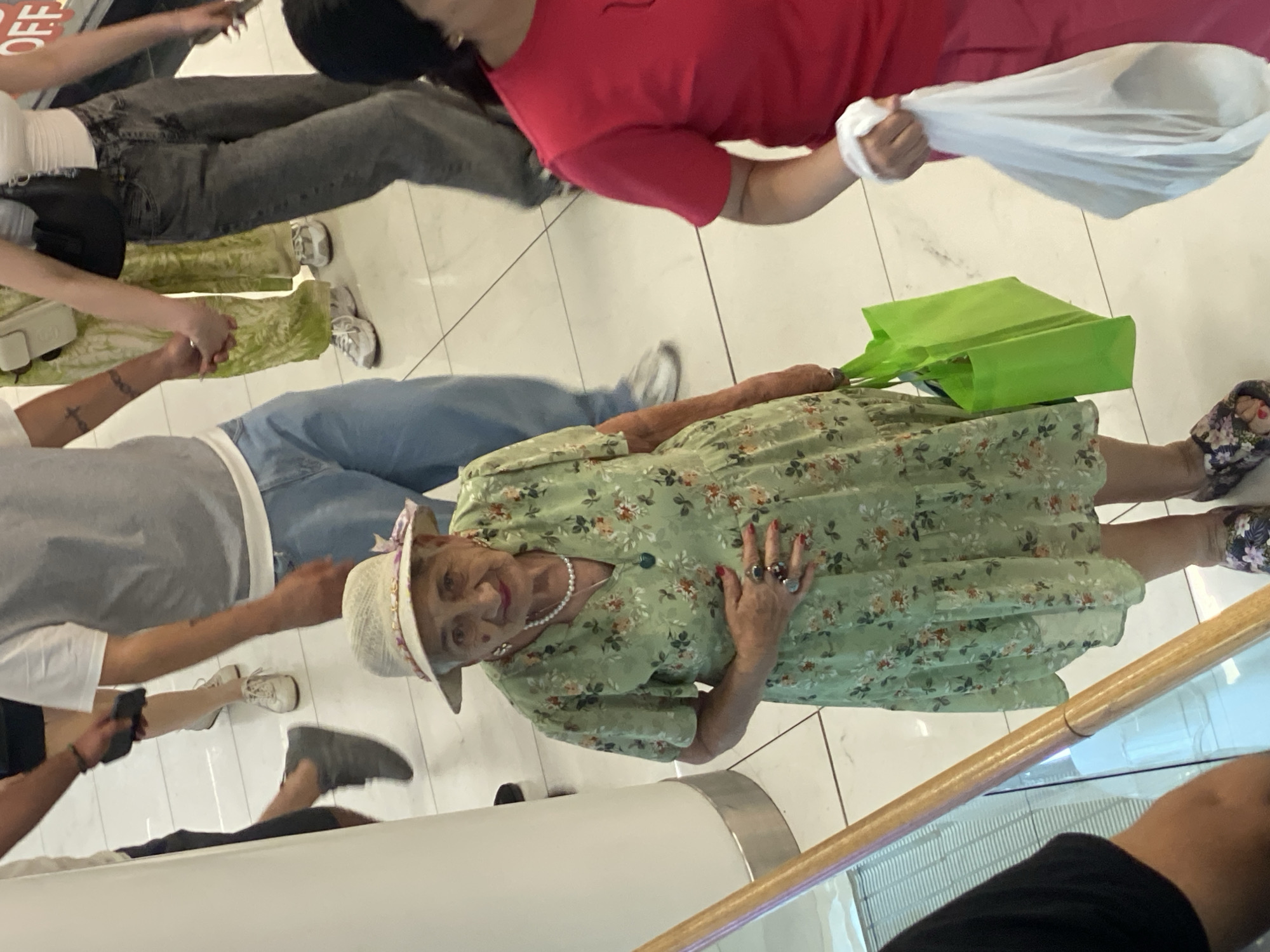An elderly woman in a light-colored, wide-brimmed hat and a green floral-print dress is pictured from the waist down, holding a bright green reusable shopping bag in her right hand. She is wearing multiple rings on her fingers and a pearl necklace. Her left hand is resting on her stomach. She is wearing colorful floral patterned slip-on shoes. The woman is standing on a tiled floor, which appears to be part of a public indoor space, possibly a shopping mall or airport, with other people in the background. The floor tiles are white with grey lines. The scene is indoors with bright lighting. The overall impression is a casual shopping environment.