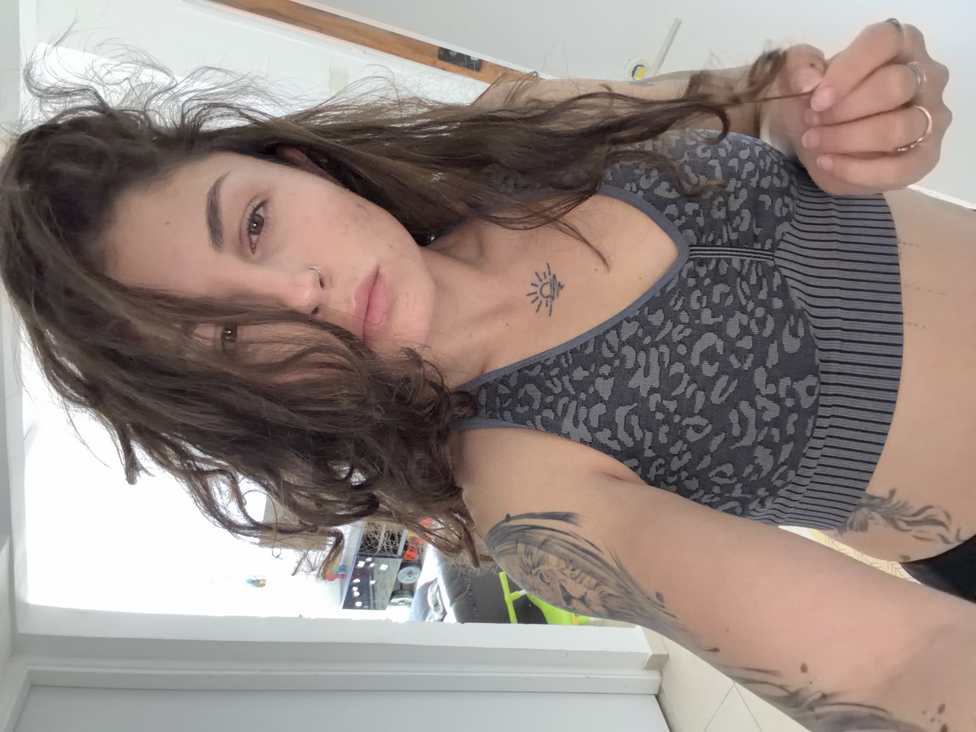 A young woman with brown hair and a nose piercing is posing for a selfie. She is wearing a gray patterned sports bra with horizontal stripes at the bottom. She has a tattoo of a sun or sunrise on her chest, and more elaborate tattoos on her arms. She is holding a strand of her hair with rings on her fingers. The background is a bright room, and through the window, some objects and vehicles are visible which could be related to Kiryat Bialik, Israel. The location is not possible to determine from this picture.