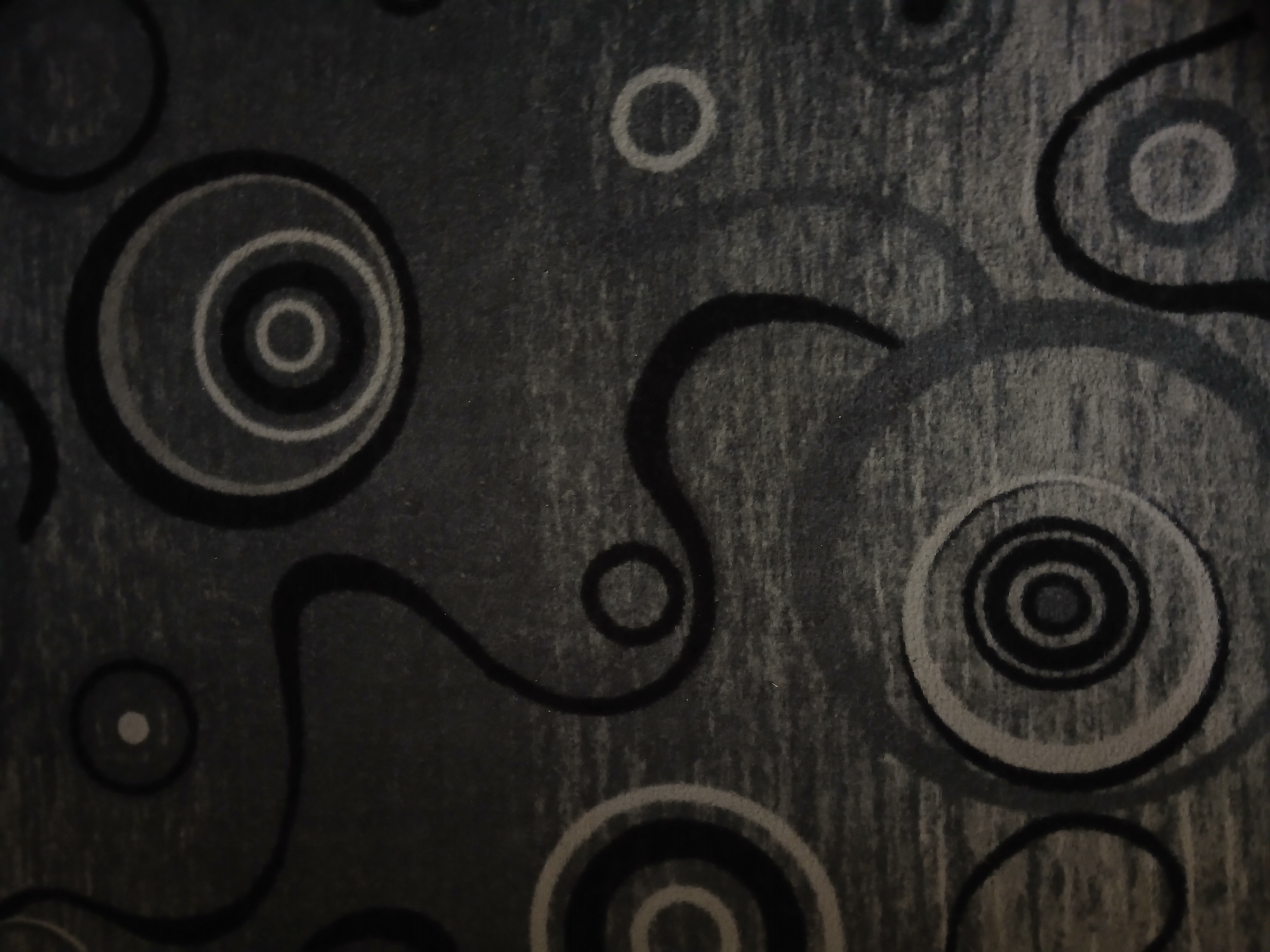 The image shows a close-up view of a dark gray carpet with a geometric pattern of concentric circles and swirling lines. The circles vary in size and are outlined in black and lighter gray. Some circles have a lighter gray or white center. The swirling lines are thick and black, creating an abstract, dynamic design across the textured surface of the carpet. There are no people, actions, or specific objects identifiable within the scene. The lighting is dim, creating shadows that emphasize the texture and patterns of the carpet. The location in Kasakh, Armenia, is not visually discernible from the image itself.