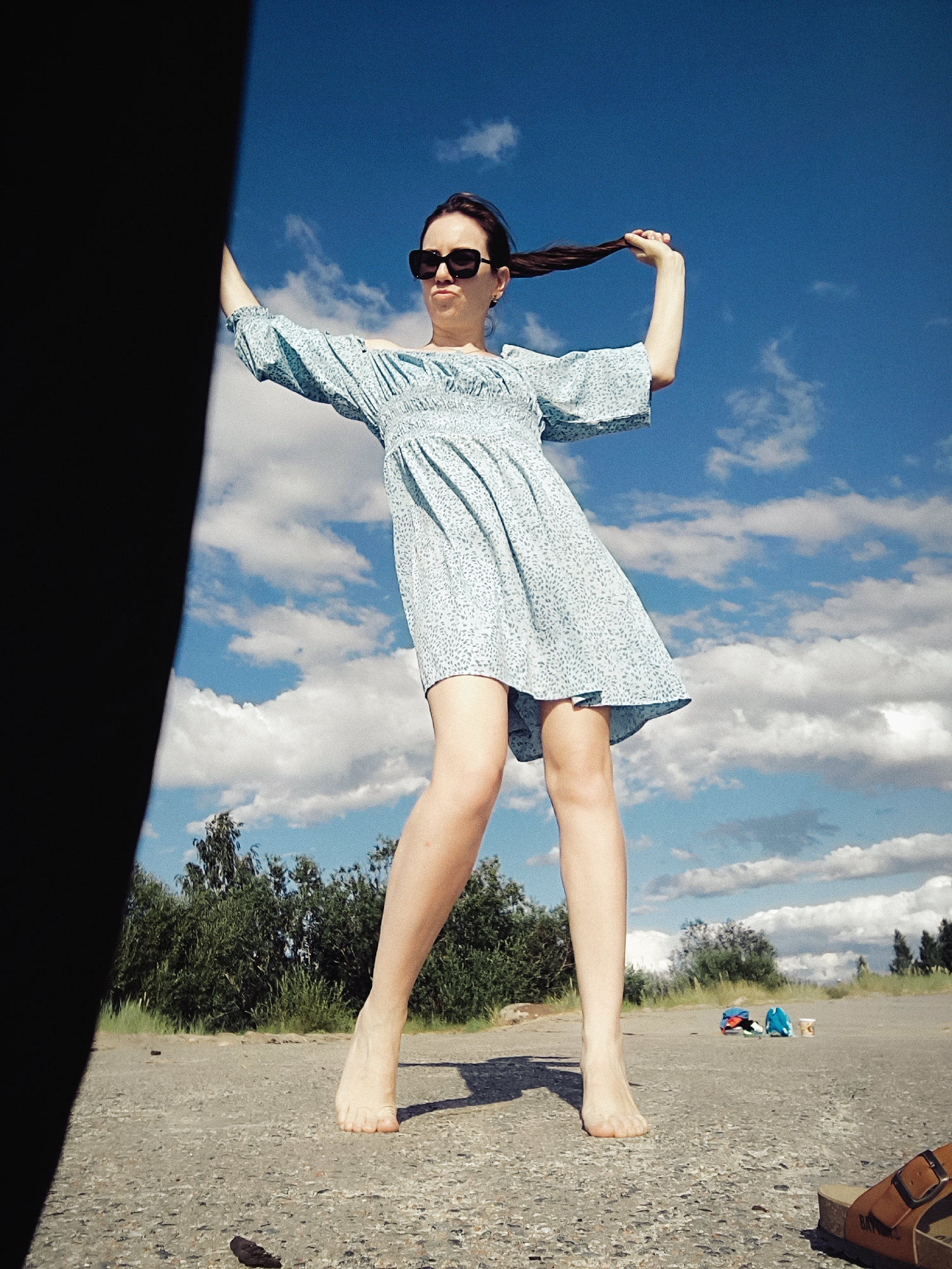 A young woman stands barefoot on a paved surface, pulling her dark hair to the side with her right hand. She is wearing a light blue, floral-patterned dress with short, puffed sleeves and black, rectangular sunglasses. Her stance suggests a playful or dramatic pose. The background is dominated by a clear blue sky with scattered white clouds. To her left, a dark, vertical object, possibly a tree trunk or shadow, partially obscures the frame. Behind her, a line of green trees and bushes stretches across the horizon. On the ground in the distance, there are a few small objects, including what appear to be bags and a cup. The lighting suggests it is daytime, possibly late afternoon, with the sun casting shadows. The location is an outdoor setting, possibly a park or a less developed area within or on the outskirts of Riga, Latvia.
