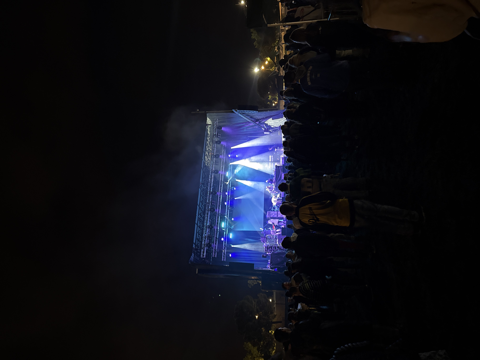 At night, an outdoor concert takes place in Vilanova i la Geltrú, Spain. A brightly lit stage displays a band performing for a crowd of people in the dark foreground. Lighting equipment is visible overhead and across the stage. Trees and streetlights are sparsely visible in the distance.